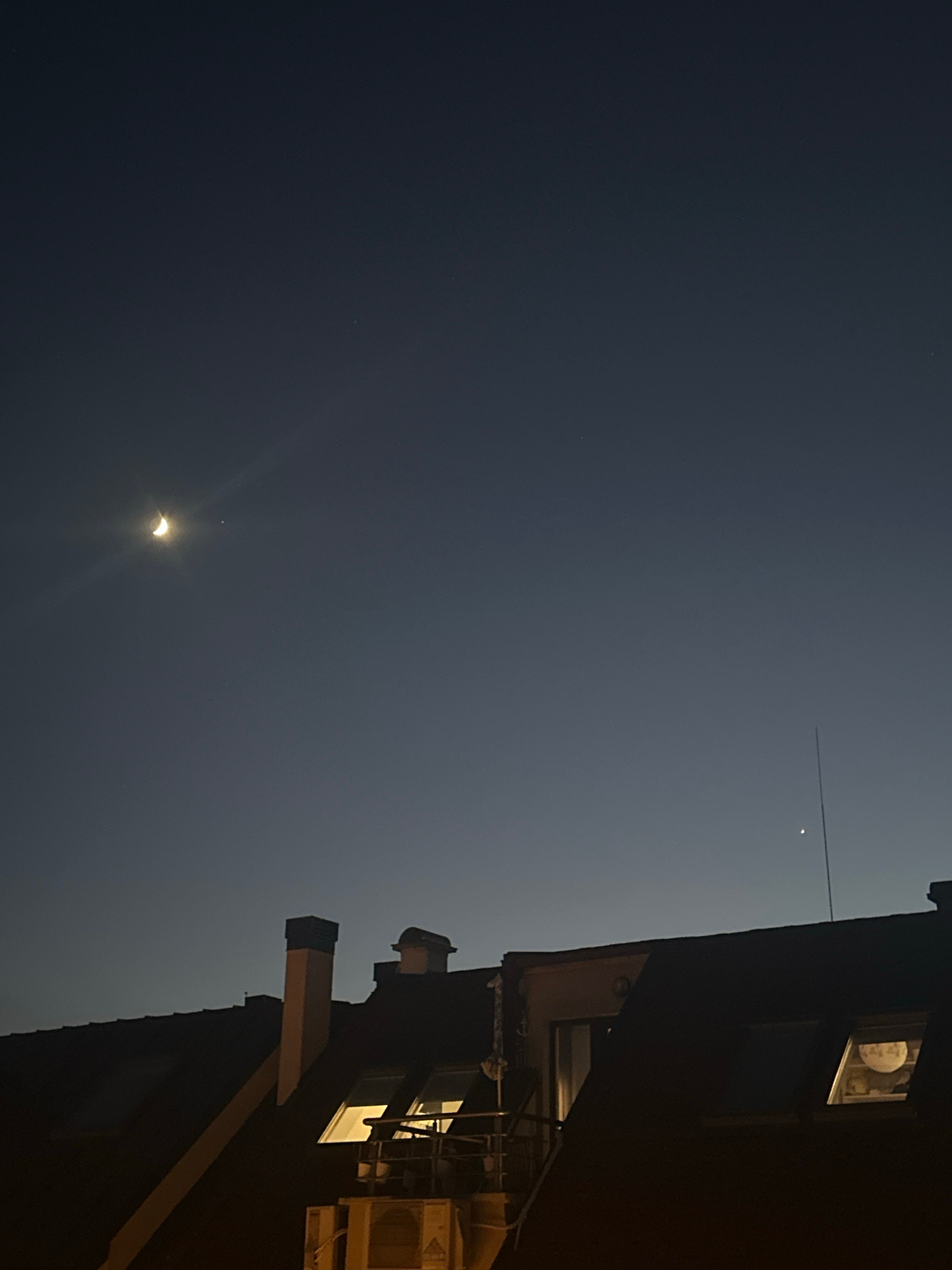 The image captures a dusky sky above the rooftops of Varna, Bulgaria. A crescent moon is visible in the upper left quadrant of the sky, emitting a soft glow. To the right of the moon, a small, bright celestial body, likely a planet, is also visible. In the lower portion of the image, the dark silhouettes of building rooftops are prominent. One building features illuminated skylight windows, suggesting interior lighting. A thin, vertical antenna extends upwards from one of the rooftops. The overall scene depicts a tranquil evening in the city.