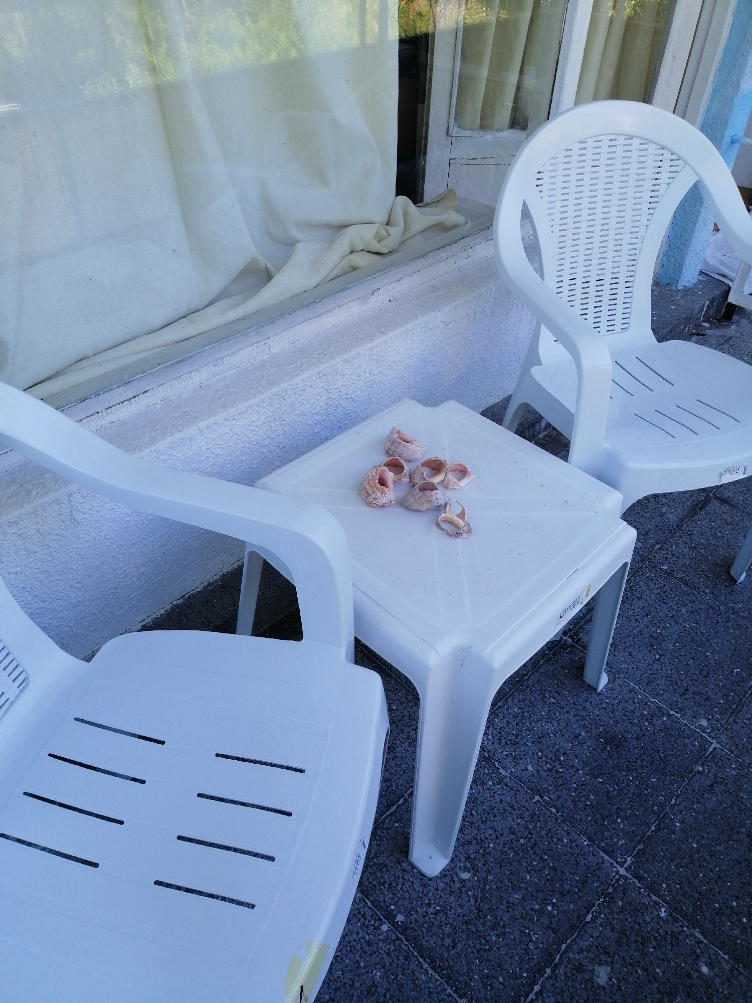 The image shows a small white plastic table with several shells on top, situated on a dark tiled floor outdoors. To the left of the table is a white plastic chair with a slatted seat and back. To the right, a portion of another white plastic chair is visible. Behind the table and chairs is a white ledge or railing, and a window with light-colored curtains is visible in the background. The overall scene suggests an outdoor seating area, possibly a balcony or patio. The image is taken from a slightly elevated angle, looking down at the table and chairs. There are no people present in the image.