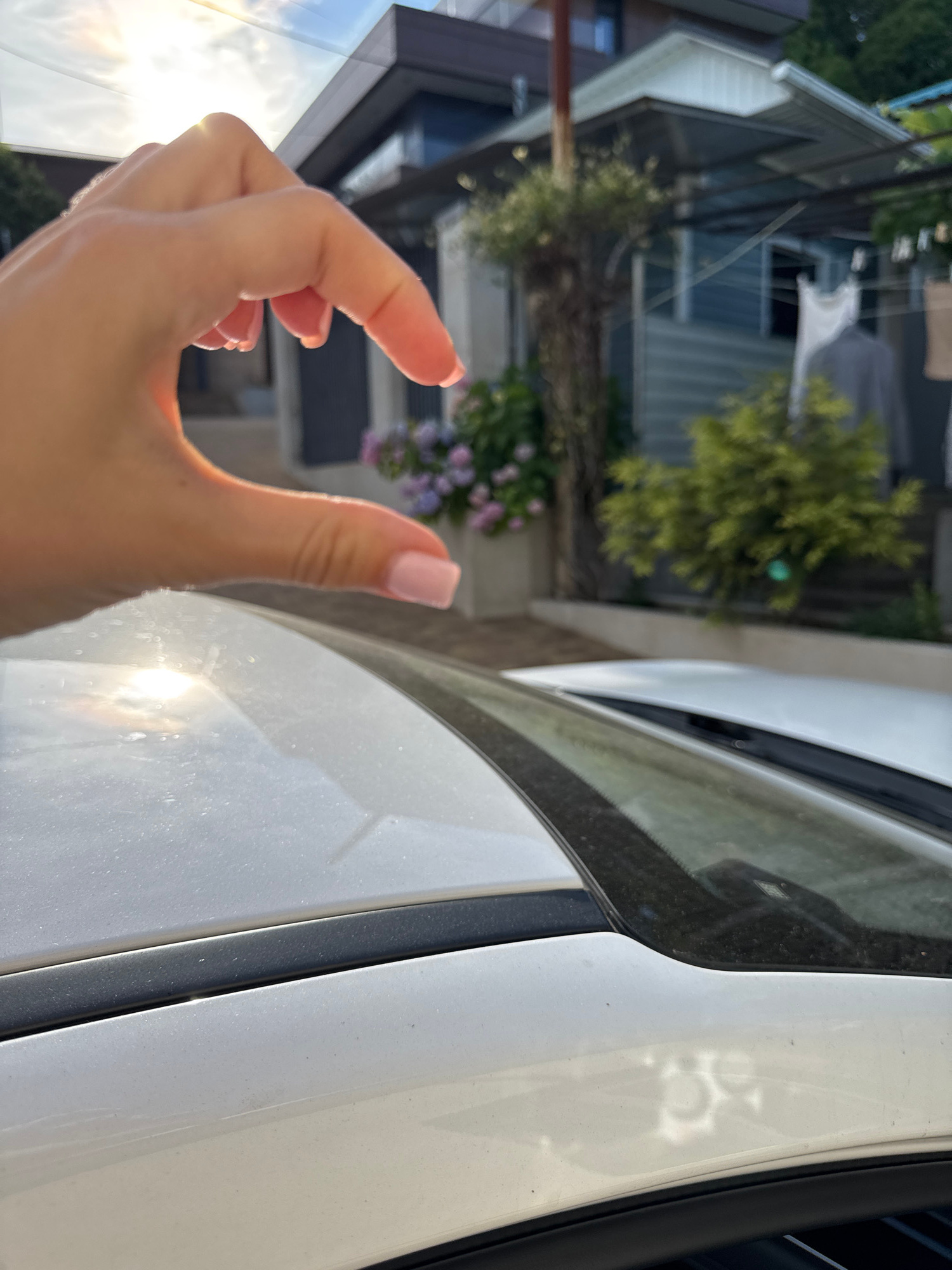 This image shows a close-up of a person's hand with painted nails, making a "pinch" gesture. The hand is positioned in front of a white vehicle, with its side and a portion of the windshield visible. The background is blurred but suggests an outdoor setting with some buildings and greenery, possibly in a residential area. The lighting indicates it is daytime, with the sun casting reflections on the car's surface. The overall mood is casual and possibly playful, given the hand gesture.
The setting is identified as Sochi, Russia. The image composition is somewhat abstract due to the close-up of the hand and the framing of the car. The focus is on the tactile gesture and the texture of the vehicle's surface.