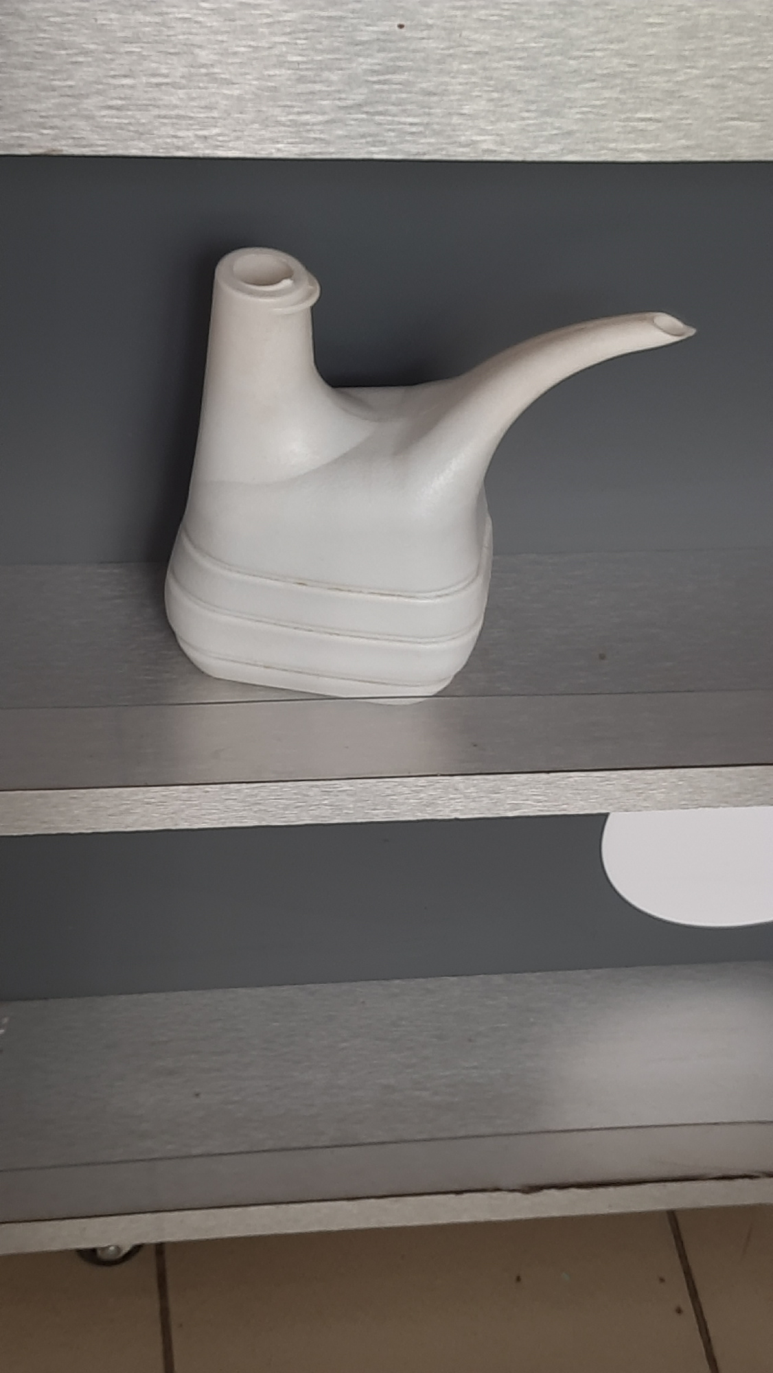 A white plastic watering can with a long spout and a rounded handle is positioned on a silver shelf. The watering can is centered in the frame and appears to be the sole object on the shelf. The shelf itself is part of a larger, dark gray cabinet or shelving unit. Above the shelf is another silver surface, possibly another shelf or the top of the unit, which has a slight texture. The background is a solid, dark gray wall. Below the shelf, the image shows another silver shelf and then the floor, which is tiled in light brown. The scene appears to be indoors, likely in a domestic or storage setting, with no discernible elements linking it to Desnogorsk, Russia.