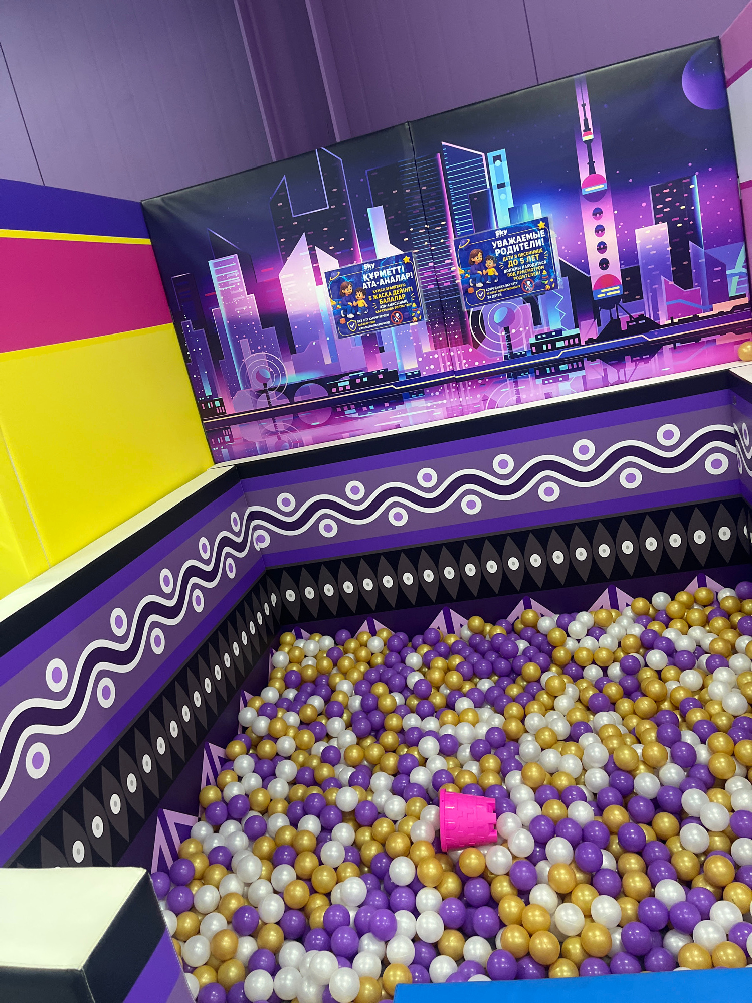 The image depicts an indoor play area, likely a ball pit, designed with a vibrant and futuristic cityscape theme. The walls are adorned with a large mural featuring stylized skyscrapers in shades of purple, pink, and blue, illuminated by neon lights. Signs with text in a non-English language, potentially Kazakh or Russian, are visible on the mural, suggesting a location in a Russian-speaking region.
The ball pit itself is filled with a large quantity of round plastic balls in three colors: purple, gold, and white. The balls are densely packed, creating a soft and colorful surface for play. A small, bright pink plastic bucket is visible amongst the balls, suggesting it might be used for scooping or carrying them.
The environment is enclosed, with patterned walls in shades of purple and black surrounding the ball pit. The upper portion of the area shows a purple ceiling with some structural elements visible. The overall impression is of a fun and engaging recreational space for children.
While the image itself does not contain explicit visual cues confirming the city of Kostanay, Kazakhstan, if this location were to be identified as such, it would represent an indoor entertainment facility within that city, catering to young children with a themed play experience.