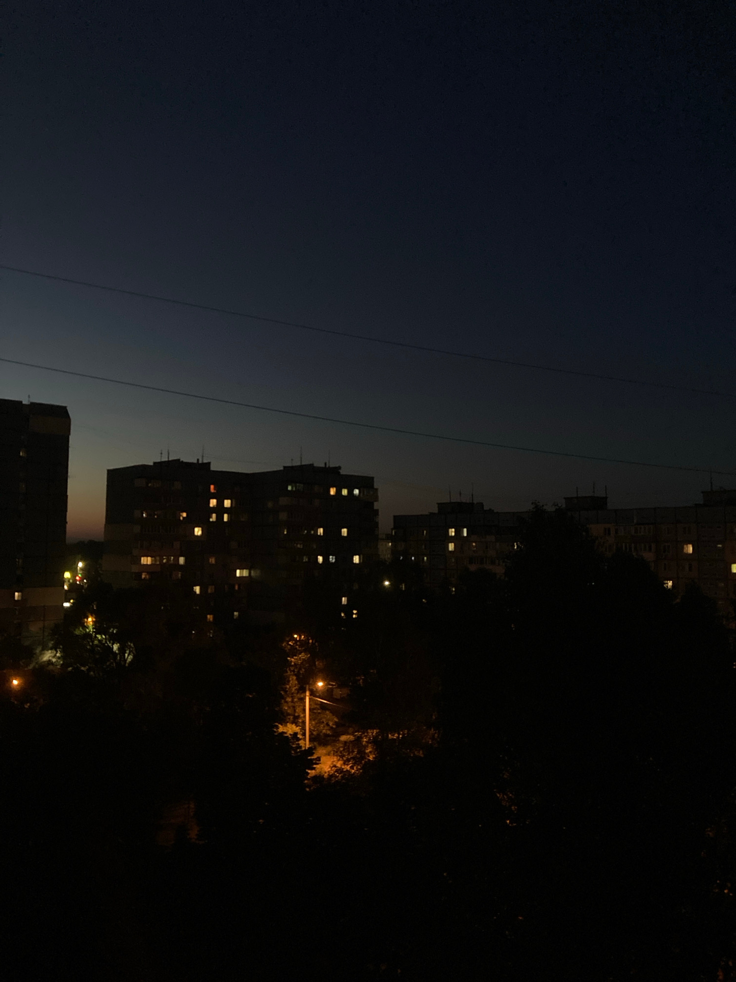 The image captures a nighttime cityscape with multiple high-rise apartment buildings illuminated by lit windows. A dark sky with a subtle gradient of twilight hues forms the background. Power lines are visible stretching horizontally across the upper portion of the frame. Silhouetted trees and foliage occupy the foreground, with streetlights casting a warm glow through the darkness, suggesting an urban residential area in Dnipro, Ukraine. The scene is devoid of any visible people or activity.