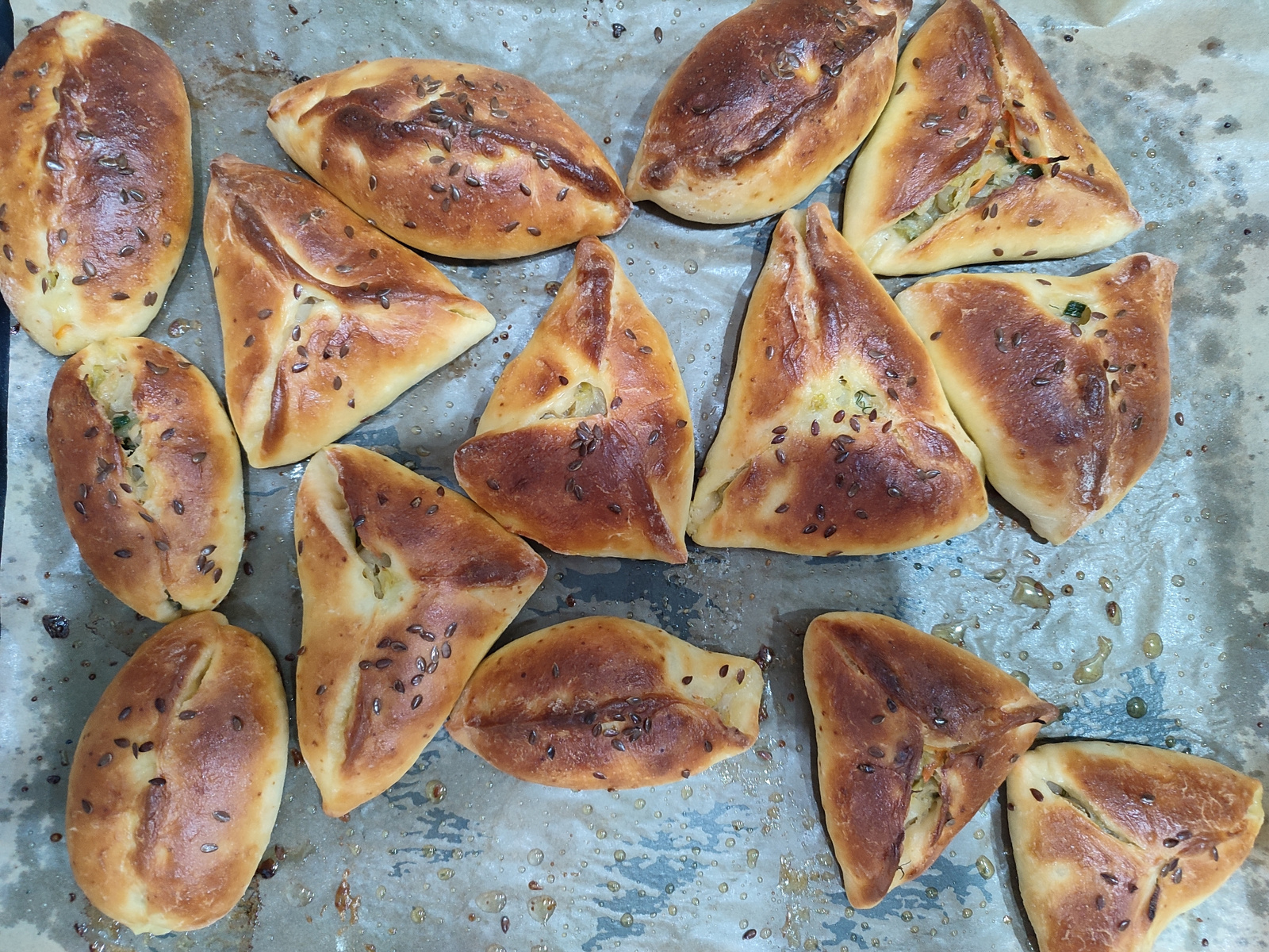 A top-down, medium shot captures a baking sheet covered in freshly baked pastries. The pastries are a mix of oval and triangular shapes, all golden brown with flecks of seeds sprinkled on top. Some of the pastries are filled, with visible hints of vegetables peeking through the openings. The baking sheet itself appears to be lined with parchment paper, which is slightly greased and has a few small droplets of oil on its surface. The scene is illuminated by overhead lighting, creating a warm, inviting glow on the food. The location is not specified, so it is unknown.