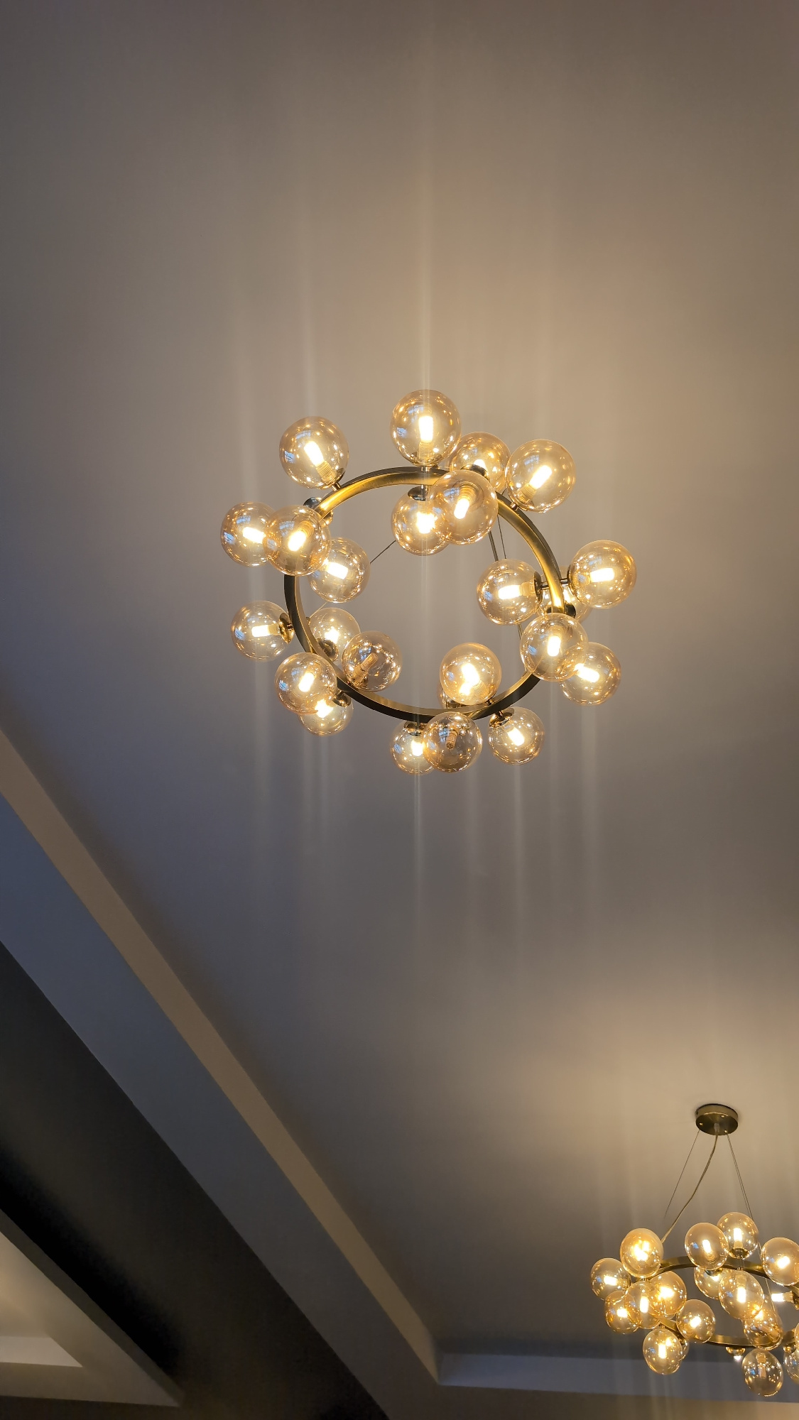 A low-angle shot captures two modern chandeliers hanging from a white ceiling. The primary chandelier, centrally positioned and larger, features a circular gold frame with numerous clear, spherical glass bulbs emitting a warm, yellow light. The bulbs are arranged in a clustered, organic manner, suggesting a dandelion or bubble-like design. The secondary chandelier, located in the lower right corner, is partially visible and appears similar in style but smaller. The ceiling has a subtle gradient of light and shadow, with vertical streaks of light emanating from the chandeliers. Architectural elements, possibly beams or ceiling molding, are visible in the lower left portion of the frame, creating a darker, angled area. The scene is static and devoid of people or explicit action. The location is Ötegen batyr, Kazakhstan.