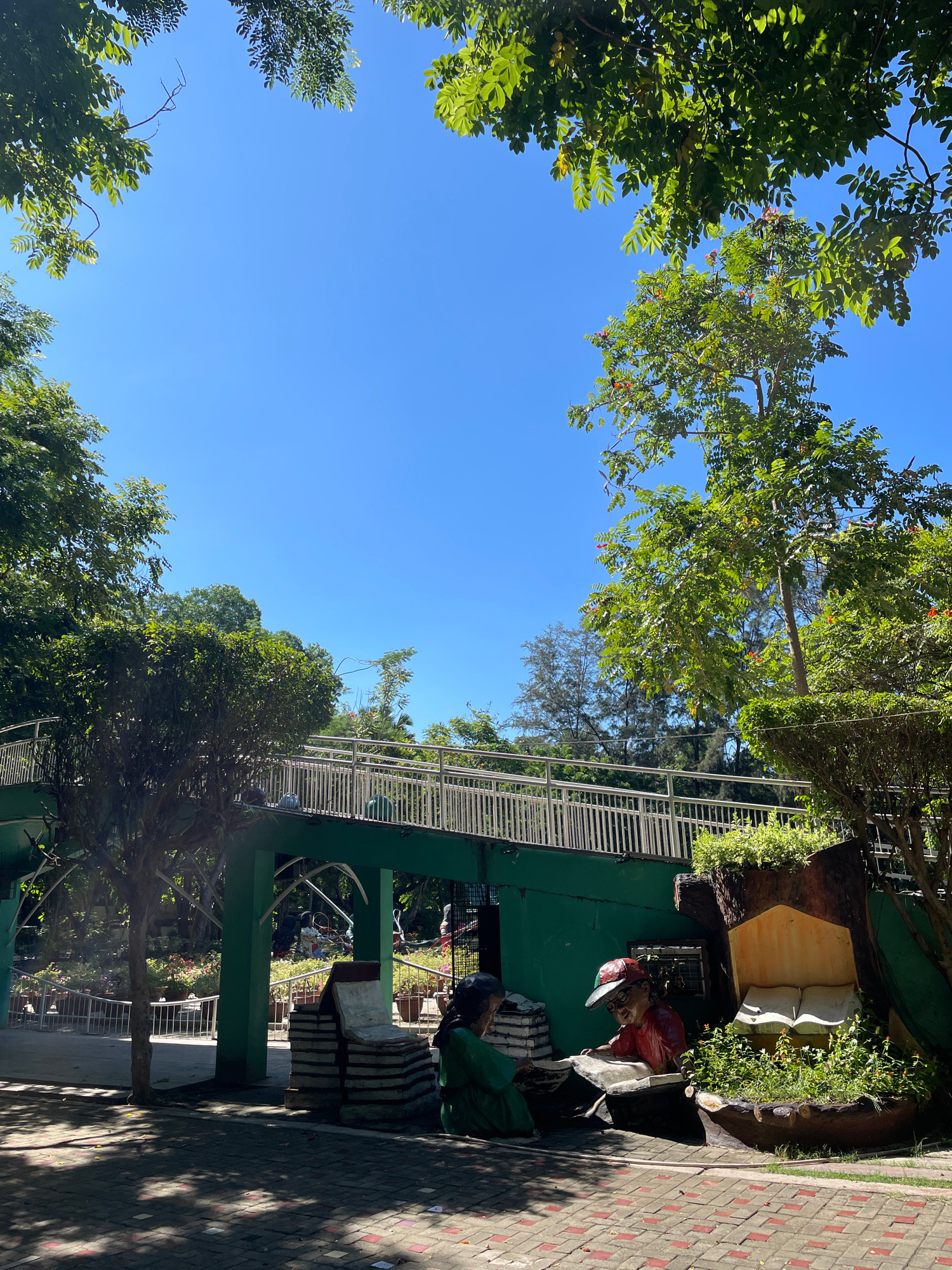 In the city of Davao, Philippines, this image captures a vibrant park scene under a clear blue sky. A green pedestrian overpass with white railings spans across the frame, connecting two sections of the park. Lush green trees with dense foliage partially frame the top and sides of the image, with sunlight dappling the paved ground below.
In the foreground, to the right, a creative installation resembling a tree trunk houses a small garden and features two child-like statues. One statue, wearing a red shirt and a baseball cap, is depicted looking down at an open book. Beside it, another statue in a green outfit is also engaged with a book, with a stack of large, book-shaped concrete objects placed behind it. The ground is paved with reddish-brown bricks and lighter-colored square tiles.
Further into the background, beyond the overpass and the statues, more greenery and potted plants are visible. The overpass appears to lead to an area with more trees and possibly other park amenities. The overall atmosphere suggests a public space designed for relaxation and recreation, with an educational element emphasized by the book-themed statues.