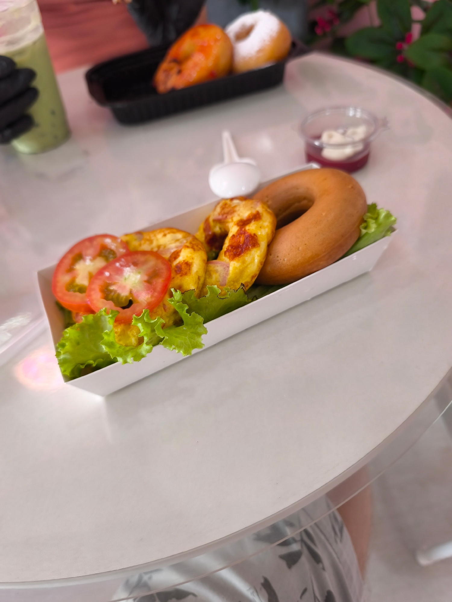 A close-up, high-angle shot shows a white disposable serving container holding a bagel sandwich. The sandwich is composed of a circular bagel, topped with two slices of tomato and lettuce. Adjacent to the bagel sandwich, in the same container, are two pieces of what appear to be cheesy scrambled eggs, one with visible browning. In the background, to the left, a hand wearing a black glove is holding a clear plastic cup with a green beverage and tapioca pearls. Further back and to the right, a small clear plastic container holds a dollop of white cream and a red jam-like substance. Behind that, a black rectangular container holds two donuts, one appears glazed and the other dusted with powdered sugar. A sprig of greenery with red berries is visible in the upper right corner. The objects are placed on a light-colored, possibly white, circular table. The scene is indoors.