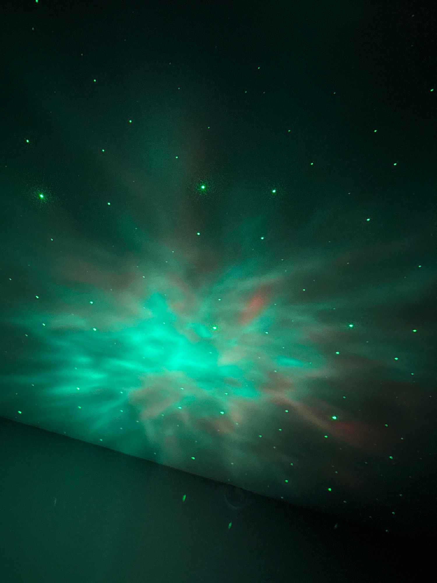 The image depicts a ceiling projection that simulates a nebula with stars. The projection features swirling clouds of green and pinkish light, with numerous small green dots scattered across the darker areas, representing stars. The projection covers a significant portion of the ceiling, creating a celestial ambiance. The lower part of the image shows a dark, solid surface, likely a wall, with some faint green light reflecting on it. The overall scene is dark, with the artificial light of the projection being the primary illumination source. There are no people or specific identifiable objects beyond the projected imagery and the surfaces it is cast upon. The location of this scene within Berezhany, Ukraine, is not visually determinable from the image itself.