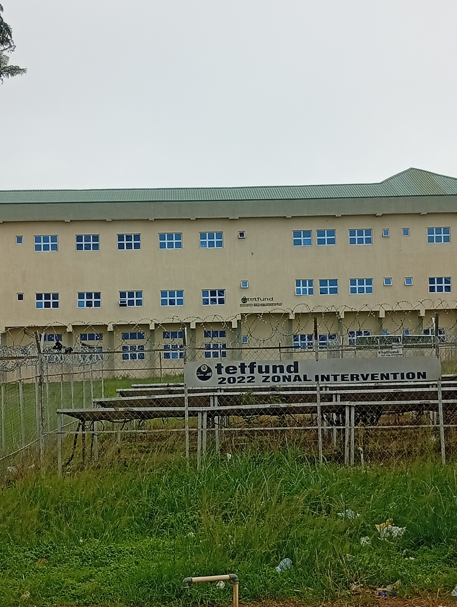 The image shows a modern, light-yellow building with a green roof, featuring many rectangular blue-framed windows. A chain-link fence topped with barbed wire runs across the foreground, separating the viewer from the building. Behind the fence, a large banner is visible, displaying text that reads "tetfund 2022 ZONAL INTERVENTION". The area behind the fence appears to be an open space with some seating structures, possibly bleachers, and overgrown green grass and weeds. The overall scene suggests an institutional or governmental facility in Wumat, Nigeria.
