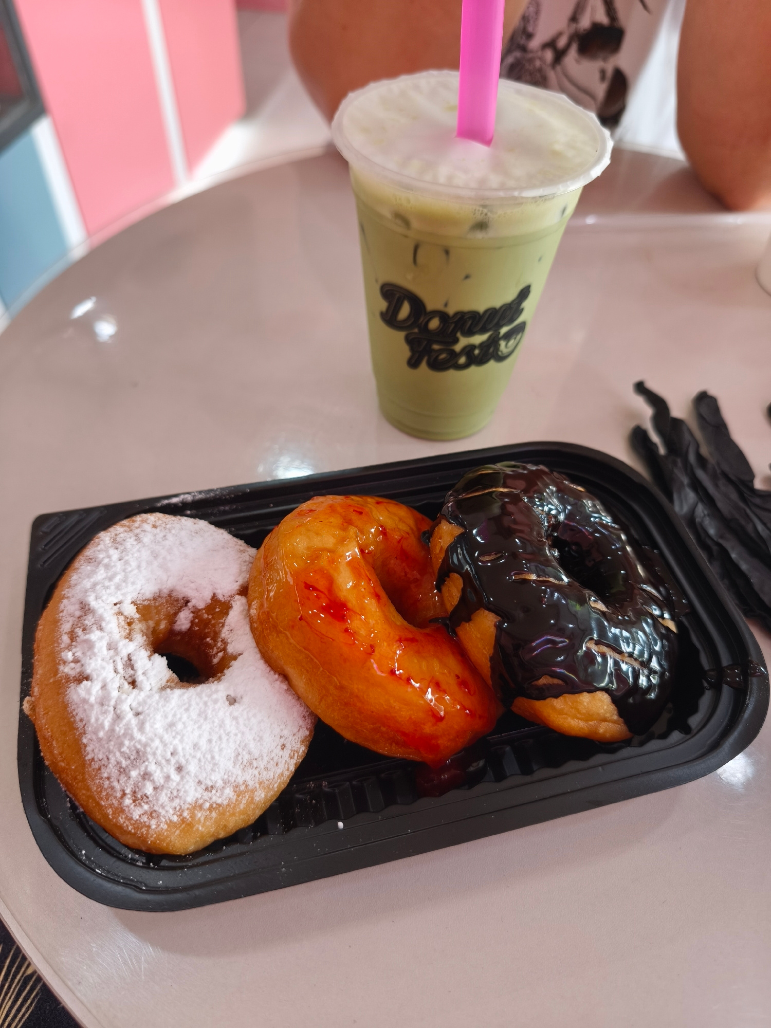 A close-up, eye-level shot shows a black plastic container holding three donuts on a light-colored table. The donut on the left is covered in powdered sugar. The middle donut is glazed with a clear orange topping and red drizzle. The donut on the right is coated in dark chocolate frosting. To the right of the donuts is a clear plastic cup with a green beverage, topped with white foam and a pink straw. The cup has a logo with the words "Donut Fest" in black lettering. In the background, parts of a person's arms and torso are visible, wearing a white t-shirt. The background also shows a blurred area with pink and blue vertical lines, possibly part of a wall or display. The scene is likely in a cafe or donut shop in Bac Nha Trang Ward, Vietnam.