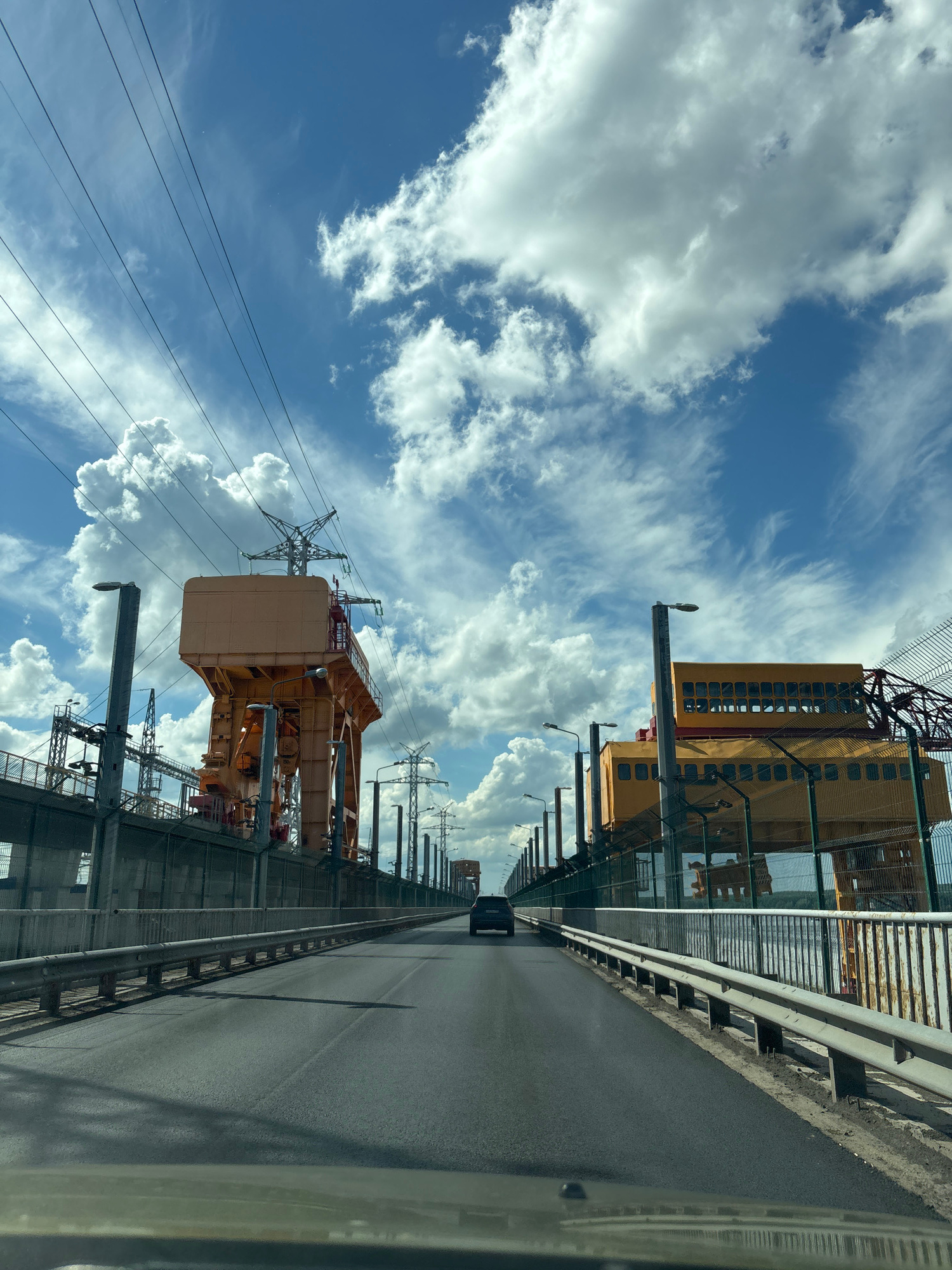 A view from a vehicle driving on a bridge, looking forward towards a dam structure. The bridge has a paved asphalt road with metal guardrails on both sides. A dark-colored car is visible driving ahead in the right lane. On either side of the bridge, there are large, industrial structures painted in yellow and orange, with visible electrical pylons and high-voltage power lines stretching across the sky. The sky is blue with large, white, fluffy cumulus clouds. The location appears to be a dam in Chaikovsky, Russia, possibly related to a hydroelectric power station. The time of day seems to be daytime with ample sunlight, indicated by the shadows on the road.
