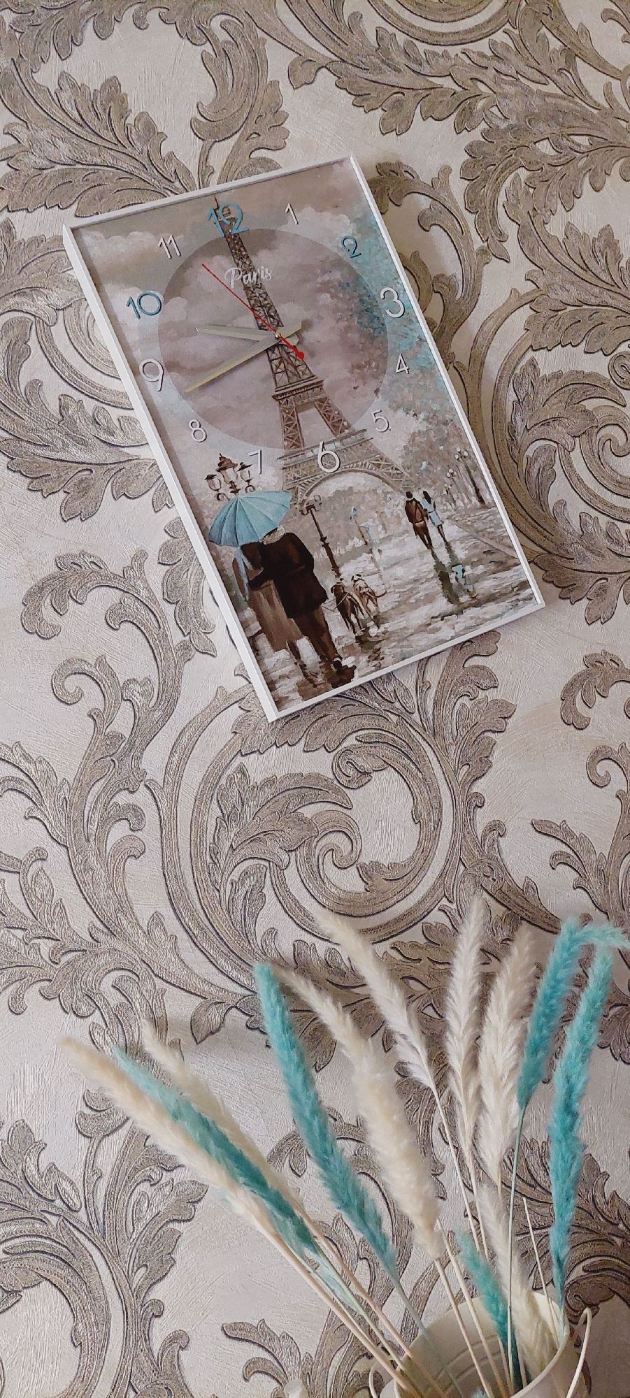 A rectangular clock with a white frame is affixed to a beige wallpaper with an ornate pattern. The clock face depicts a watercolor painting of the Eiffel Tower in Paris, with a couple walking under an umbrella with dogs in the foreground, on a wet cobblestone street. The clock's hands are red and light gray, with the hour hand pointing to approximately 3 and the minute hand pointing to approximately 10. In the lower right corner of the image, a bouquet of dried pampas grass and other decorative stems, some colored teal, are arranged in a white vase. The scene is lit by overhead lighting, casting soft shadows.