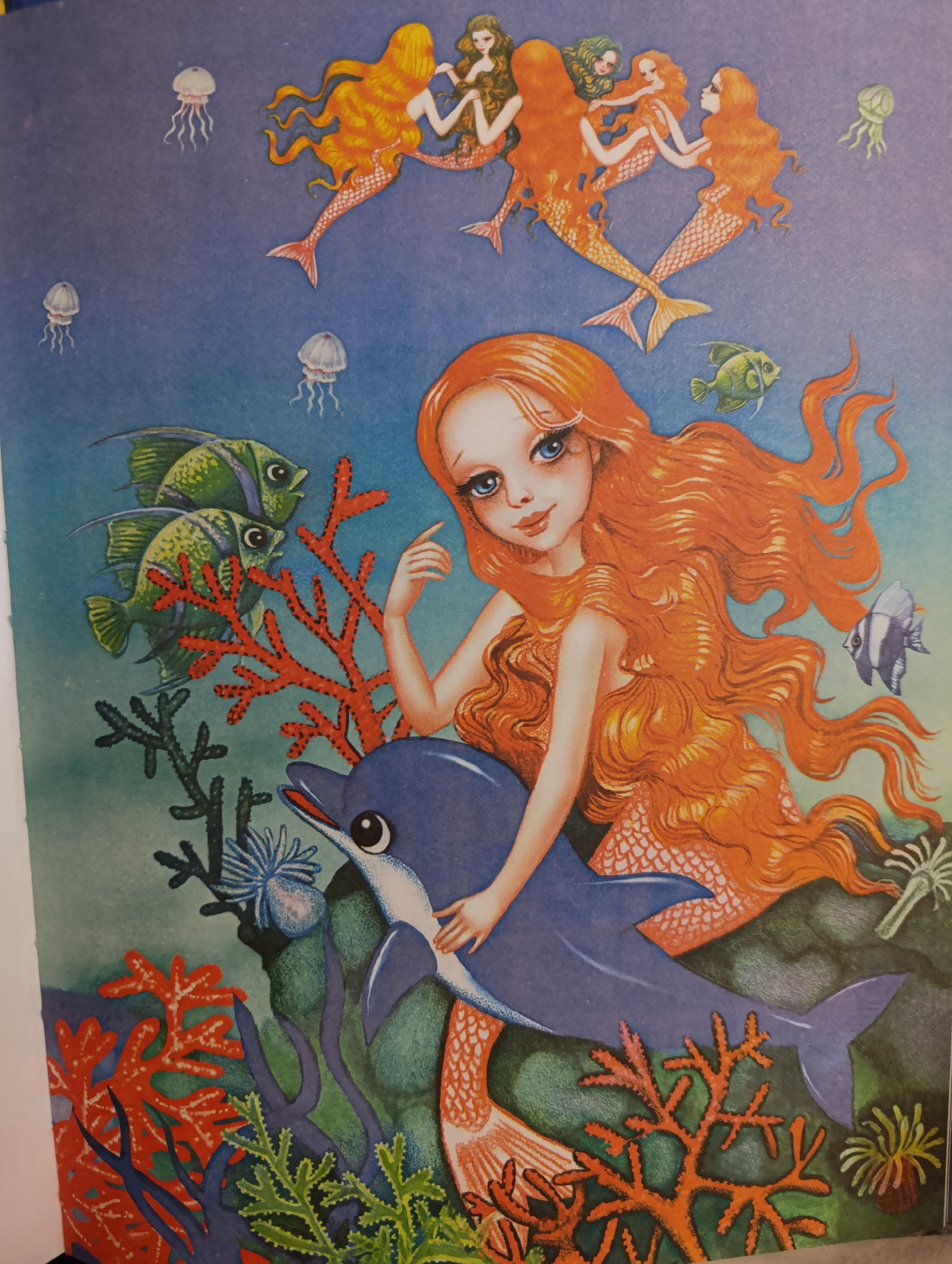 The image depicts an underwater scene rendered in a whimsical, illustrative style. In the foreground, a young mermaid with bright blue eyes and long, flowing orange hair is shown riding a blue dolphin. She has a fishtail with orange scales. The dolphin's mouth is slightly open, and its eye is large and black. The mermaid's left hand rests on the dolphin's back, and her right hand is extended upwards.
In the background, several other mermaids with orange hair and fishtails are shown dancing or interacting in the water. They are positioned in the upper portion of the image. Scattered throughout the blue water are several small, translucent jellyfish.
The underwater environment is populated with various marine elements. Two green fish with blue stripes are swimming near the left side of the image. Red coral formations are prominent, with some dark green and blue plant-like structures also present. A small striped fish is visible to the right of the main mermaid. The overall color palette is vibrant, with blues, greens, and oranges dominating the scene.
The image is a static illustration and does not depict any specific location within a city.
