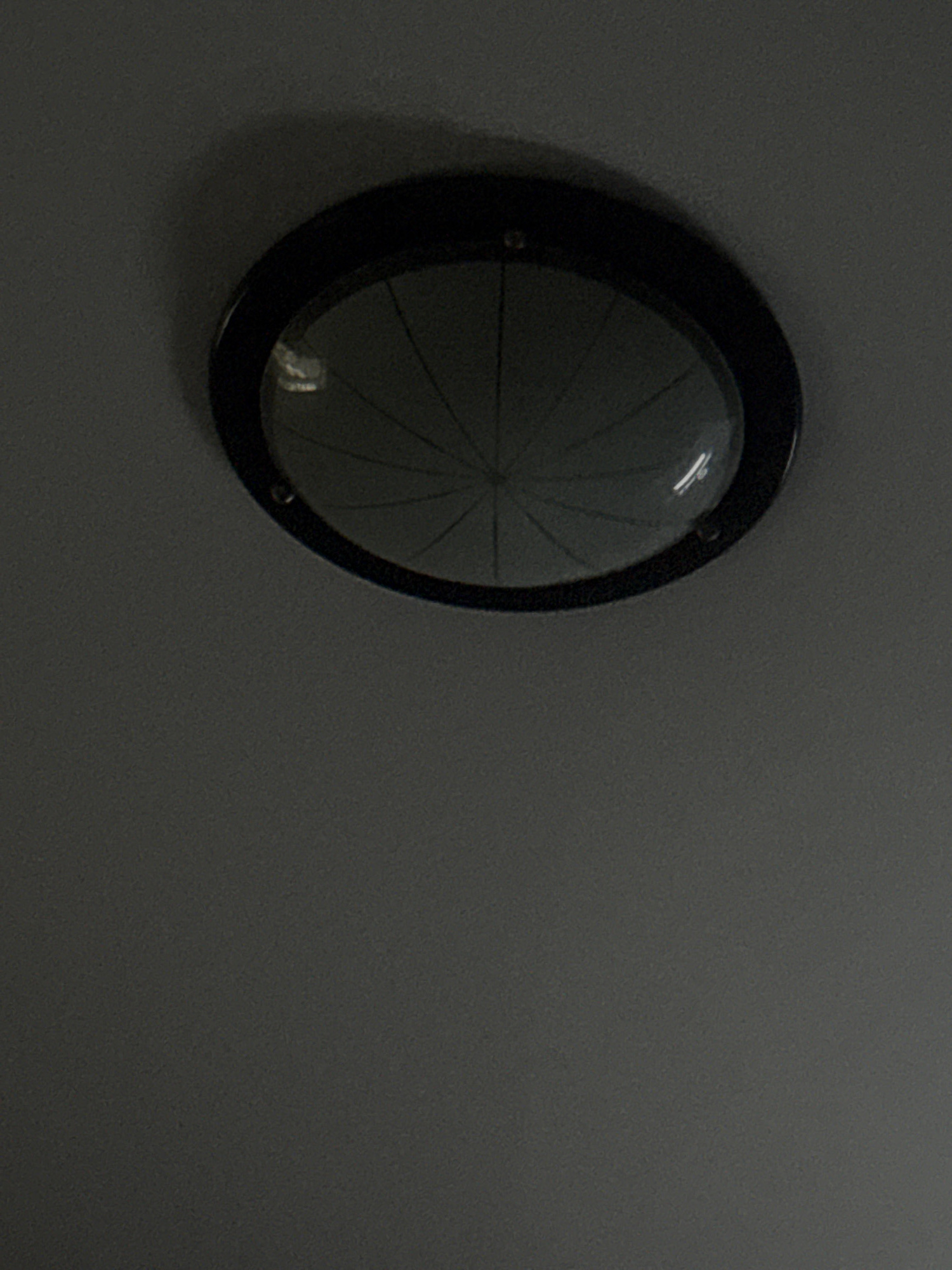 The image shows a ceiling light fixture with a black frame and a clear, patterned glass cover. The light fixture is centrally located in the upper portion of the frame against a dark gray, textured ceiling. The glass cover has a radial pattern, with lines emanating from the center. There are reflections visible on the glass, suggesting an interior environment. The image is dark, with the light fixture being the primary source of illumination in the scene. There are no people or other objects visible. The image is taken from a low angle, looking up at the ceiling.