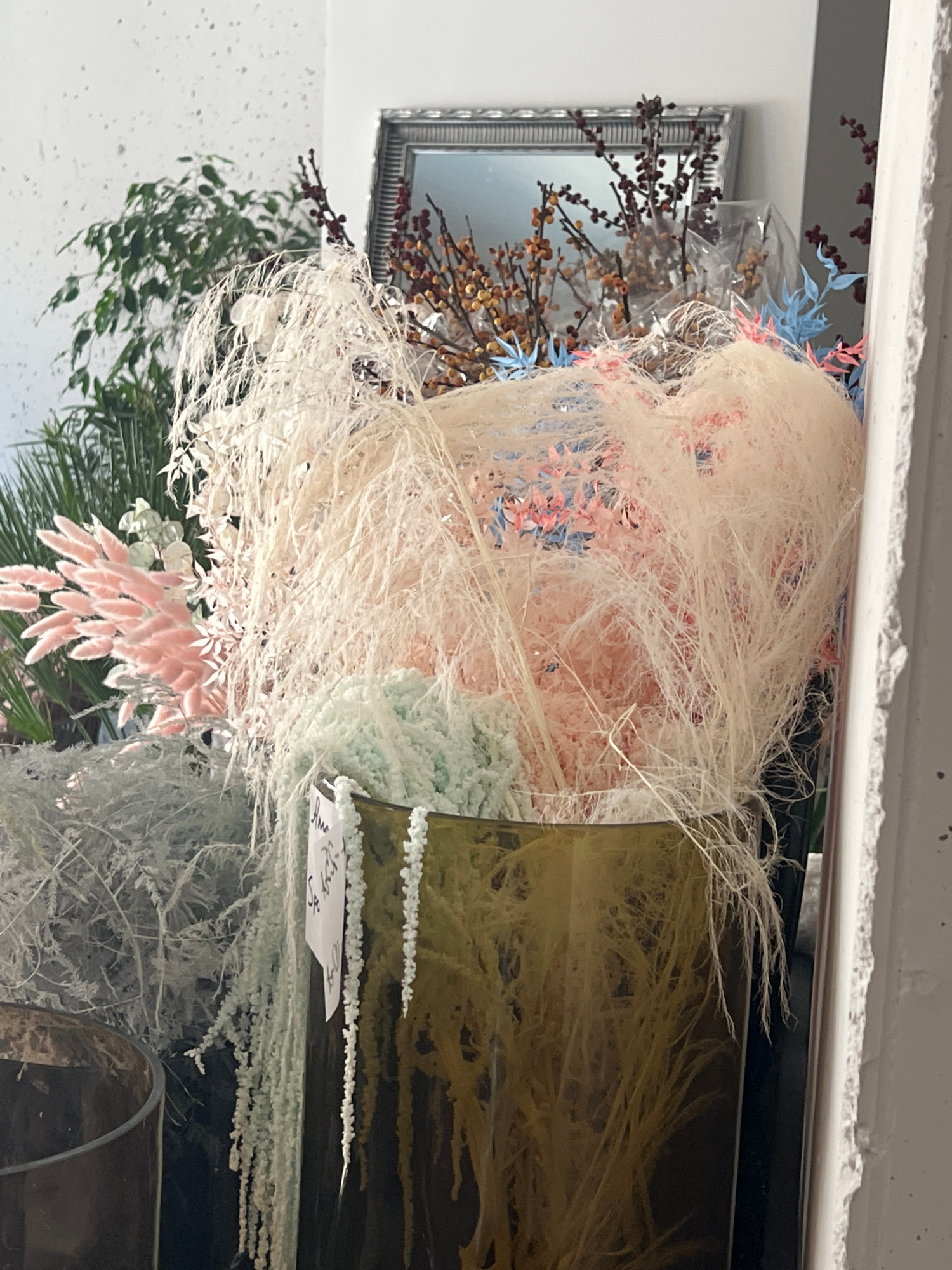This is a close-up, vertically oriented photograph of a decorative arrangement of dried flowers and grasses. The subjects are various dried botanical elements, including what appears to be pampas grass or a similar feathery grass in shades of cream and beige, delicate pink feathery flowers, and sprigs of small brown berries on branches. The arrangement is presented in what seems to be a glass vase or container, partially visible on the right side of the frame, which has a greenish-brown hue. The overall aesthetic is textural and natural, with a focus on delicate, wispy forms.
In the background, a silver-framed mirror is visible, reflecting some of the decorative elements and a wall. There are also hints of blue and pink artificial leaves or flowers incorporated into the arrangement. The lighting appears to be soft, suggesting an indoor setting, possibly a shop or a home interior. The overall mood conveyed by the arrangement is calm and perhaps somewhat whimsical due to the ethereal quality of the dried materials.
On the side of the glass container, a small, white tag with handwritten text is affixed. The text is partially obscured and difficult to read clearly but appears to be in a script, possibly Polish. The letters "Krakow, PL" are visible, confirming the location context. There are no people or clear indicators of a specific time of day or weather conditions. The focus is entirely on the artistic presentation of the dried floral arrangement.
