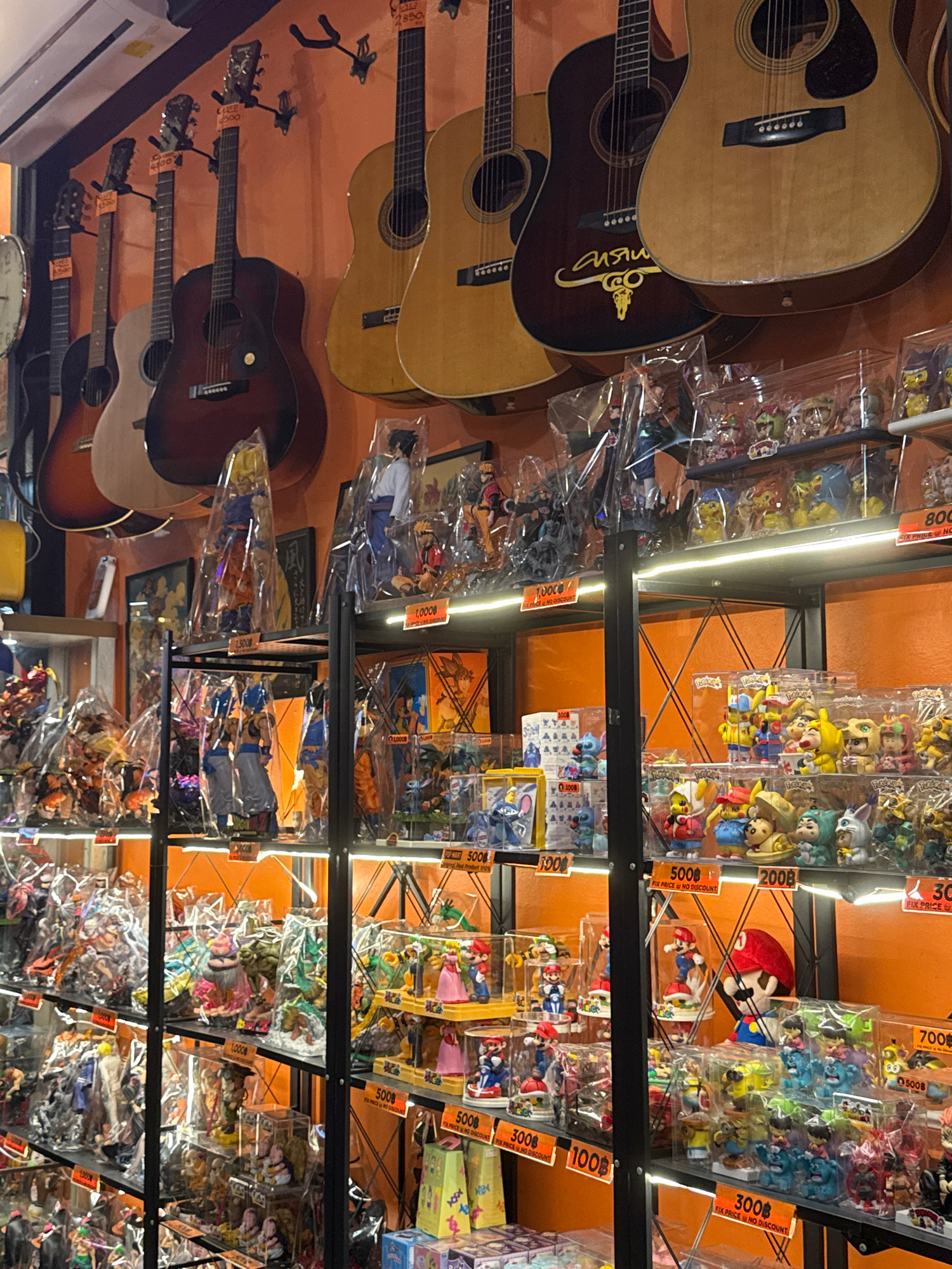 The image shows a retail store with shelves displaying various items, primarily acoustic guitars and collectible figurines. The guitars are hung on the walls and displayed on stands, with several visible at different angles. The figurines, many of which appear to be anime or manga-inspired characters, are packaged in plastic and arranged on shelves.
Several price tags are visible in the image, with prices written in both numbers and Thai Baht currency (฿). Some tags also include text like "FIX PRICE NO DISCOUNT" and "ADS". The setting appears to be indoors, likely a market or a specialty store, given the eclectic mix of merchandise. The lighting suggests it could be daytime or under artificial lighting within the store. The overall impression is of a bustling marketplace where musical instruments and collectibles are sold. There are no people clearly visible in the image, suggesting the photo was taken when the store was not crowded or the individuals were out of frame.