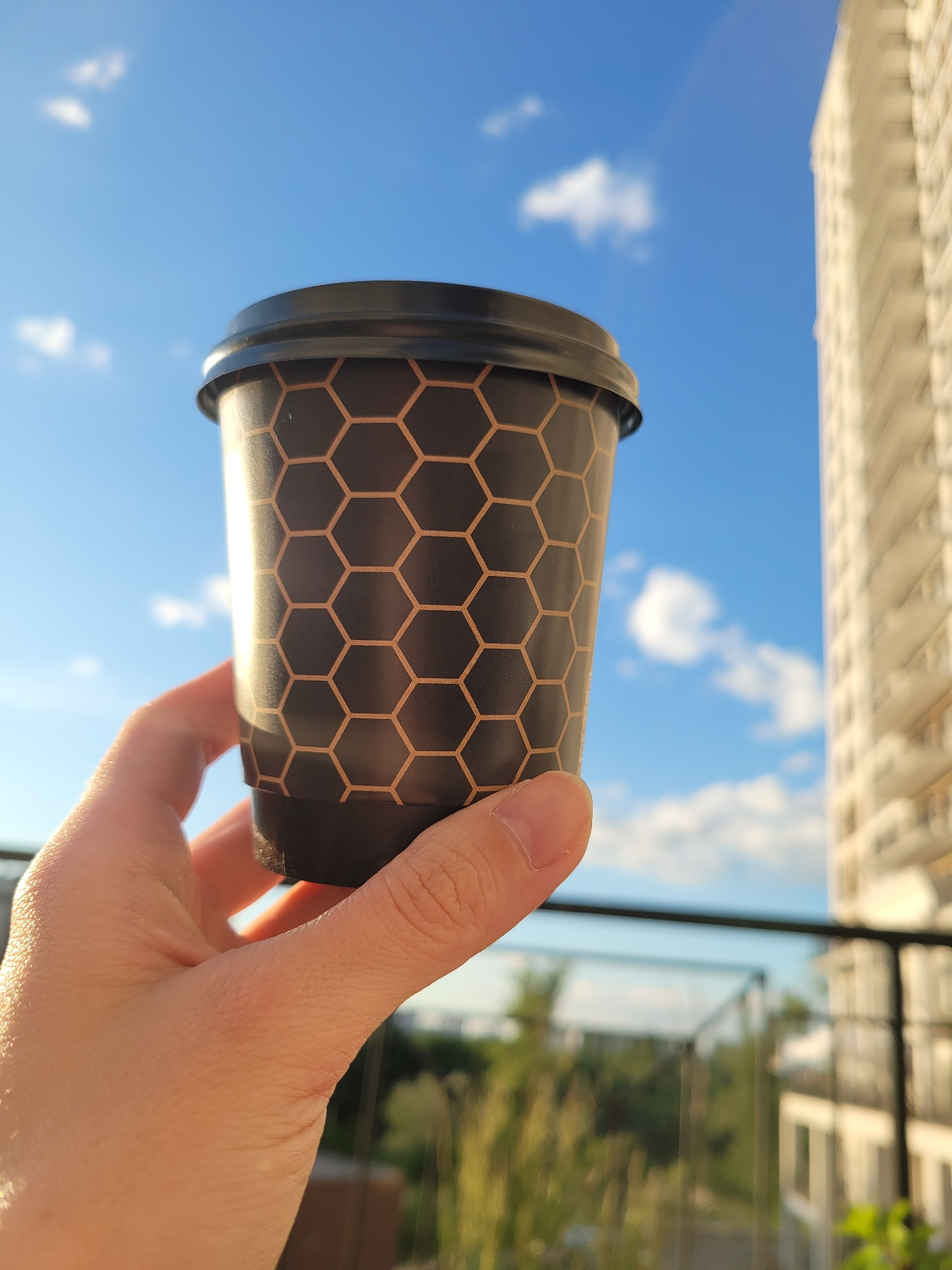 A hand holds a black paper coffee cup with a hexagonal pattern against a bright blue sky with scattered white clouds. In the background, to the right, is a blurred view of a modern apartment building. The scene appears to be set outdoors, possibly on a balcony, with a railing visible in the foreground. The image has a shallow depth of field, with the cup and hand in sharp focus and the background out of focus. The lighting suggests it is daytime, possibly late afternoon due to the warm glow on the hand and cup. The location is the city of Kyiv, Ukraine.