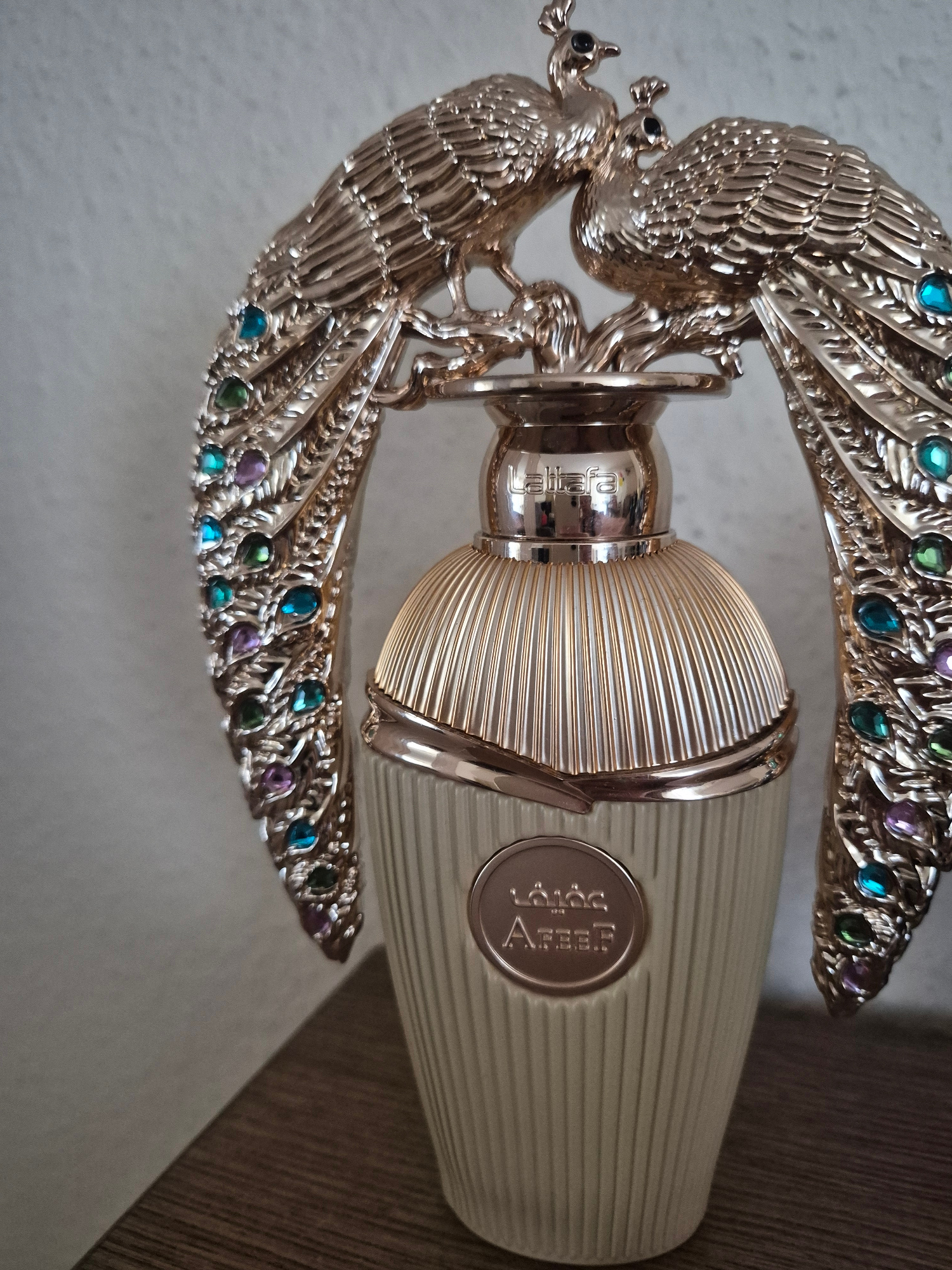 A gold-colored perfume bottle is displayed against a light-colored, textured background. The bottle is adorned with a decorative gold embellishment featuring two peacocks with outstretched wings. The peacocks' wings are decorated with colorful, faceted gems in shades of blue, green, and purple. The bottle itself has a ribbed texture and features a circular emblem with Arabic script and the word "Abeer" in English. The bottle is placed on a dark wooden surface.