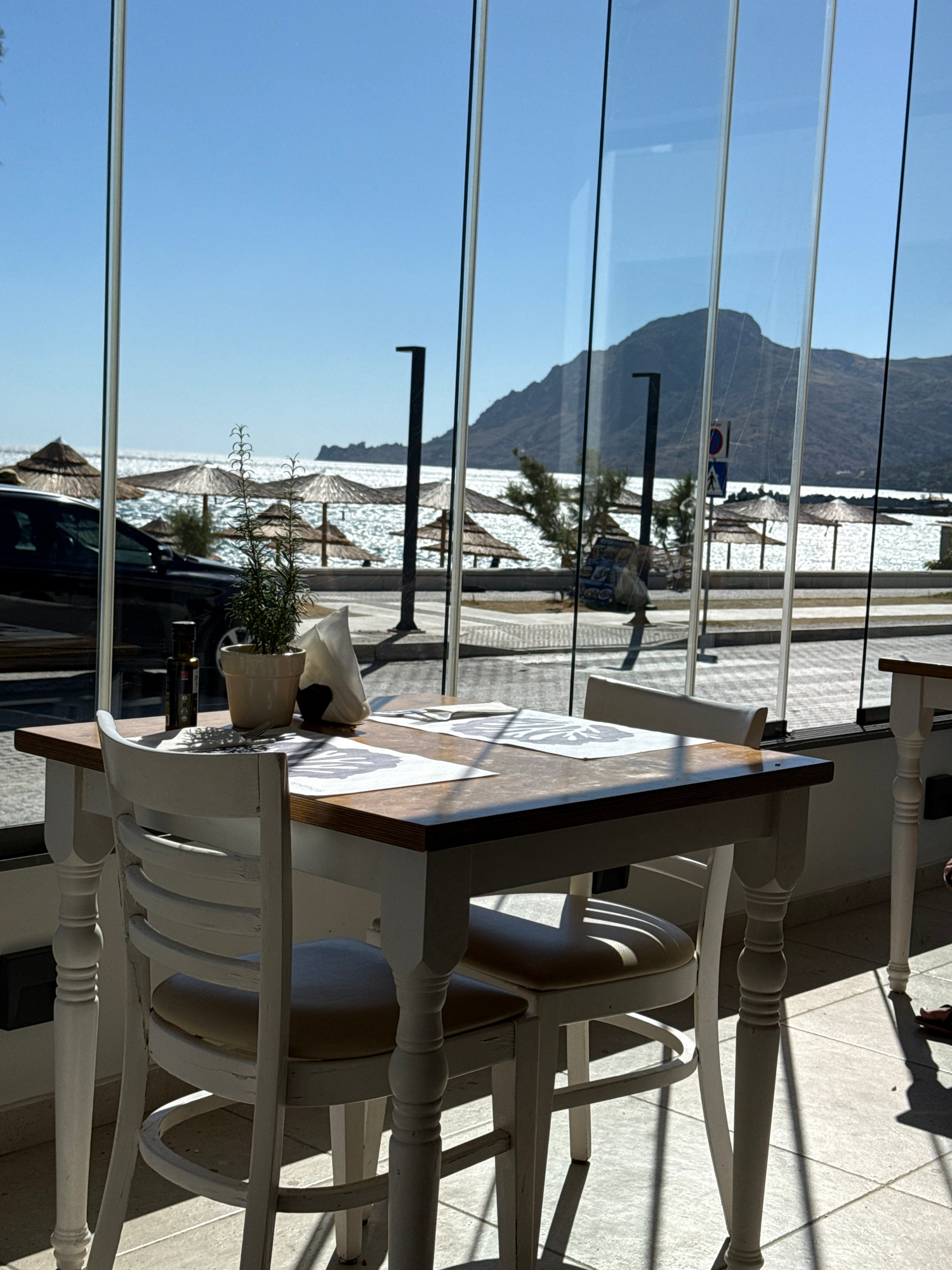 The image shows a table and chairs set up inside a building, with a large window looking out onto a beach scene. The table is made of wood with a white base, and it has two white chairs with light-colored cushions. On the table, there is a potted plant, a bottle of olive oil, and a napkin holder. Outside the window, there are several thatched umbrellas on the beach, and the sea is visible in the background. A large hill or mountain is also visible in the distance, behind the sea. The sun is shining brightly, casting shadows on the floor inside the building. There are no people visible in the foreground, but a person's feet and leg are visible on the right side of the image in the background. The overall impression is of a restaurant or cafe with a view of the sea.