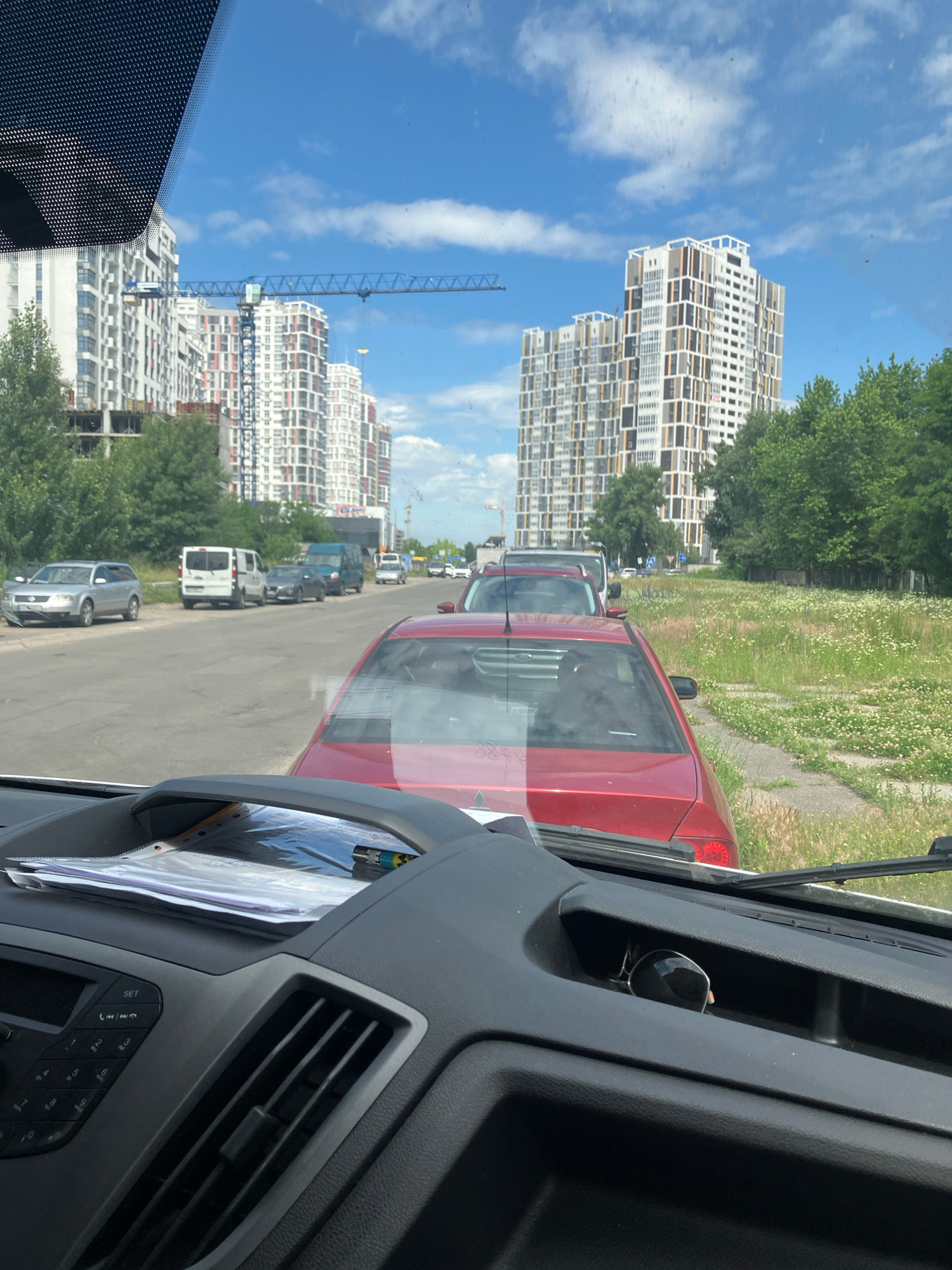 The image shows a view from inside a vehicle, looking out through the windshield onto a city street in Kyiv, Ukraine. The street is lined with parked cars and a few vehicles are in motion. On the left side of the street, a silver station wagon is parked, followed by a white van and a dark gray sedan. Further down the street, a blue van and other vehicles are visible. On the right side of the street, a red sedan is parked, with its rear facing the camera. Beyond the street, there are residential buildings under construction, with cranes in the background. The sky is blue with scattered clouds, and green trees and grassy areas are present. The interior of the vehicle is visible in the foreground, with a dashboard, air vents, and papers on a holder. Sunglasses are resting in a compartment on the dashboard. The scene appears to be a typical urban environment on a sunny day.
