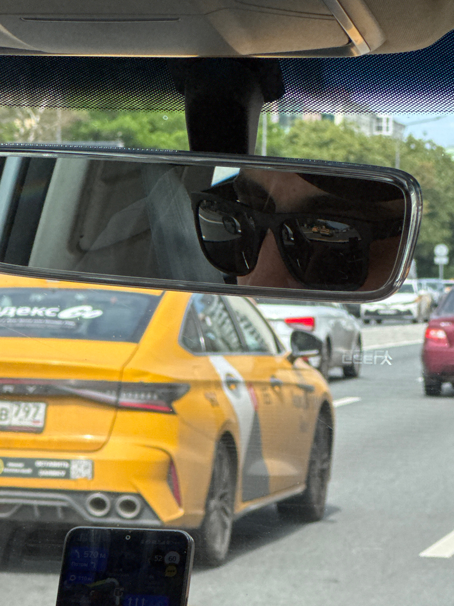 The image shows a view from inside a vehicle, looking forward through the windshield and at the rearview mirror. In the rearview mirror, a person wearing sunglasses is reflected, looking at the road ahead. The road appears to be a multi-lane street in a city, with several other cars visible. The primary subject is a yellow taxi with Russian license plates, which is in front of the viewing vehicle. The taxi has markings that suggest it is from Yandex Taxi. In the foreground, a smartphone is visible displaying a navigation app with route information. The scene suggests traffic on a sunny day in Moscow, Russia, likely during daylight hours.