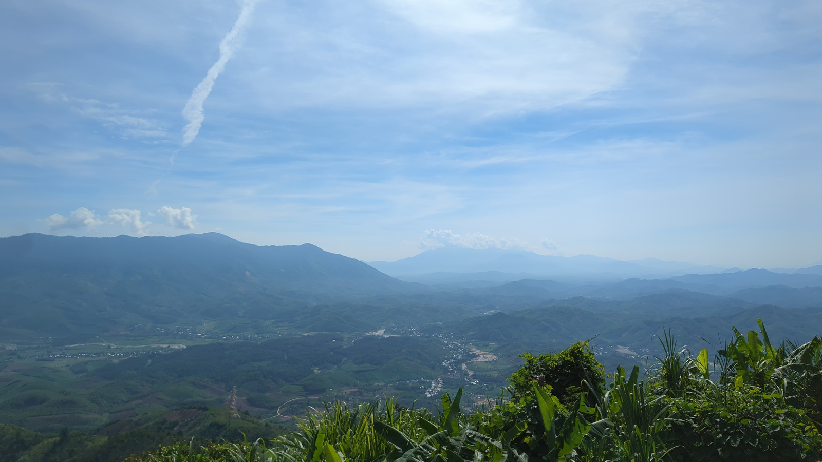 This is a wide-angle, high-angle panoramic photograph taken from a vantage point overlooking a valley near Tay Khanh Vinh, Vietnam. The foreground is dominated by lush green vegetation, including tall grass and broad-leafed plants. Beyond the vegetation, a vast valley unfolds, filled with rolling hills covered in a patchwork of green agricultural fields and scattered human settlements. A river or waterway meanders through the valley floor, reflecting the sky. In the distance, successive layers of hazy blue mountains recede into the horizon, their forms softened by atmospheric perspective. The sky above is a clear blue with scattered white clouds and a prominent contrail stretching diagonally across the frame. The overall scene depicts a rural landscape under a bright, clear sky.
