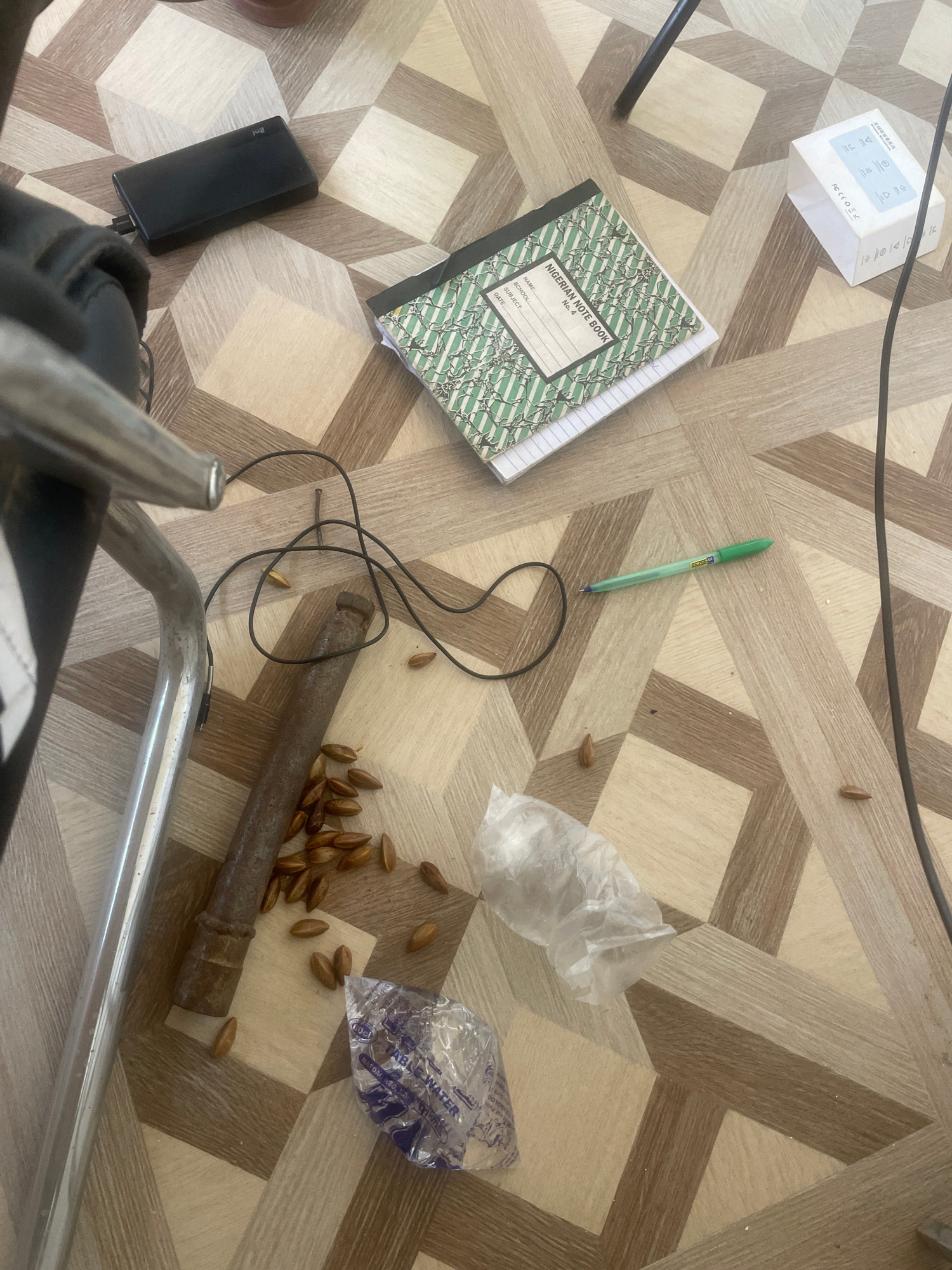The image depicts a floor with a geometric tiling pattern composed of light and dark brown wooden planks. Several objects are scattered across the floor, suggesting a casual or disarranged setting.
In the upper left quadrant, a black rectangular power bank is visible, with a cable extending from it. Adjacent to the power bank, a green pen lies on the floor.
A "Nigerian Note Book" with a green and white striped cover is situated in the center of the image, placed on top of a plain white notebook. The note book has fields for Name, School, and Date.
In the lower half of the image, a collection of small, brown, oval-shaped objects, possibly seeds or nuts, are scattered. Among these is a rusty, cylindrical metal object, likely a piece of pipe or metal rod. A thin black wire is also visible, looping around some of the seeds and near the metal object.
Two plastic bags are present: one is crumpled and translucent, appearing empty or containing a small amount of material. The other is a clear plastic bag with blue lettering, partially visible, that appears to be a packet of "TABLE WATER".
The lighting in the image is moderate, casting soft shadows and highlighting the texture of the floor and objects. The scene appears to be indoors, on a floor that could be found in a residential or communal area. The mention of "Jos, Nigeria" in the prompt suggests a potential geographical context for the scene, although no specific landmarks or indicators of this location are present within the image itself. The overall impression is one of everyday objects left on the floor.