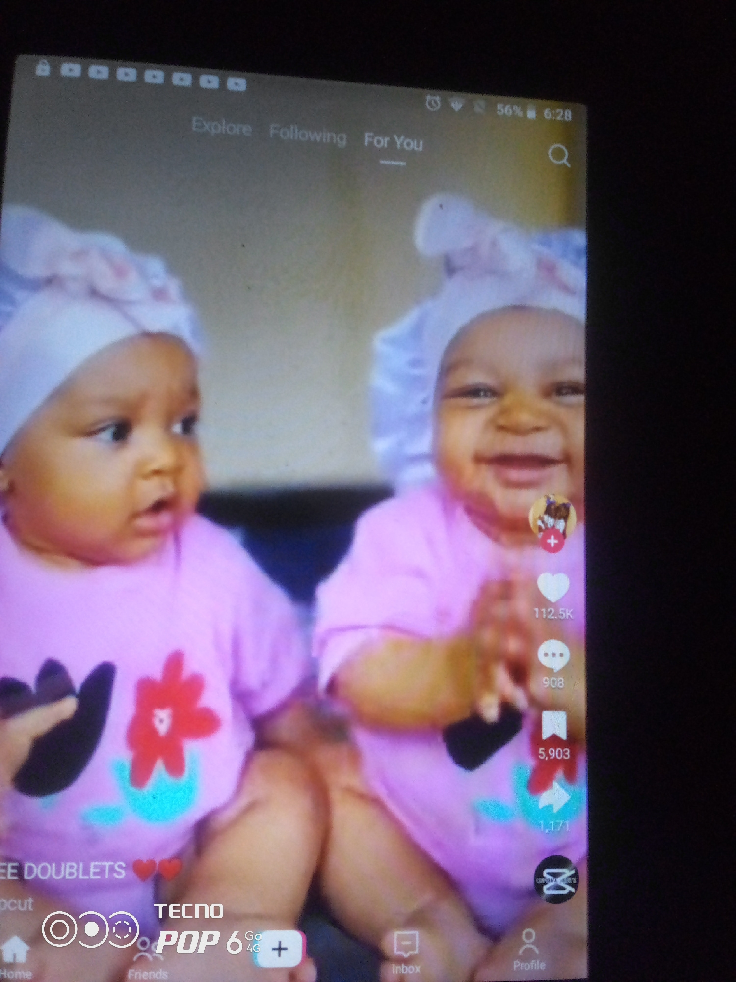 The image is a screenshot from a mobile device, likely a social media application like TikTok, showing two infants wearing pink shirts and bonnets. The infants are positioned close together, with the one on the left looking to the side with an open mouth, and the one on the right smiling at the camera. Overlayed on the image are various interface elements of the app, including icons for likes, comments, shares, and profiles, as well as text indicating engagement metrics like "112.5K" likes and "5,903" shares. A "TECNO POP 6" logo is visible at the bottom, suggesting the device used to capture the screenshot. The background is a neutral, indoor setting. There is no information in the image that could identify its location within Howayi, Nigeria.