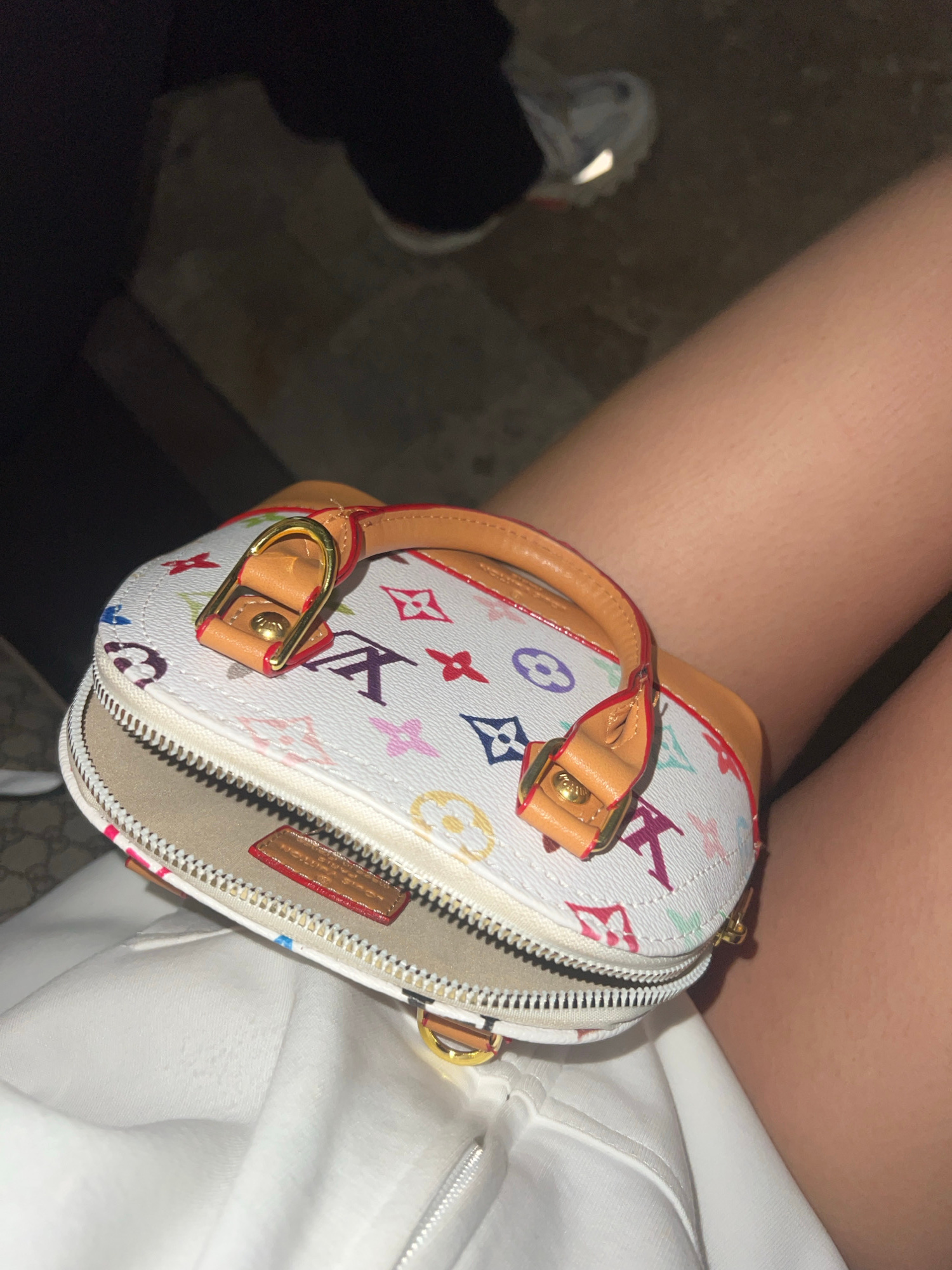 A small, white, multi-colored Louis Vuitton Speedy Nano bag with brown leather trim and gold hardware is displayed on a person's lap. The bag is partially open, revealing a tan interior. The person is wearing a white hoodie or sweatshirt. In the background, out of focus, is a dark area and what appears to be a shoe with reflective detailing. The scene is dimly lit, suggesting it may be nighttime or indoors. There is no information about the location in Zlatograd, Bulgaria.