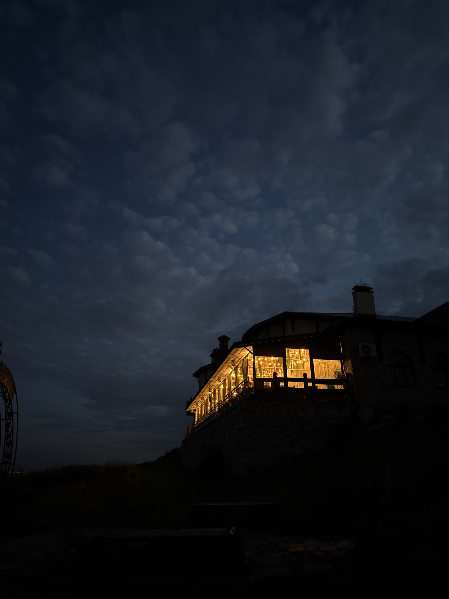 This low-angle, vertical, nighttime photograph captures a rustic, stone-faced building with a long, brightly lit, enclosed veranda extending along its side. The veranda is illuminated by numerous warm-toned lights, possibly string lights, creating a cozy and inviting atmosphere within. The building itself appears to be constructed with natural stone and has a dark, sloped roof with a visible chimney. A dark, possibly wrought-iron, decorative archway is partially visible on the left edge of the frame, suggesting an entrance or garden feature. The foreground is in deep shadow, with only hints of vegetation and possibly some low seating. The background is dominated by a dark, cloudy sky, with subtle variations in tone and texture within the clouds. The overall impression is one of a secluded, atmospheric evening scene, possibly a restaurant or guesthouse, situated in a natural or semi-natural environment. There are no people or animals visible in the frame. The location is identified as Yelabuga, Russia, suggesting a rural or historical setting within that city.