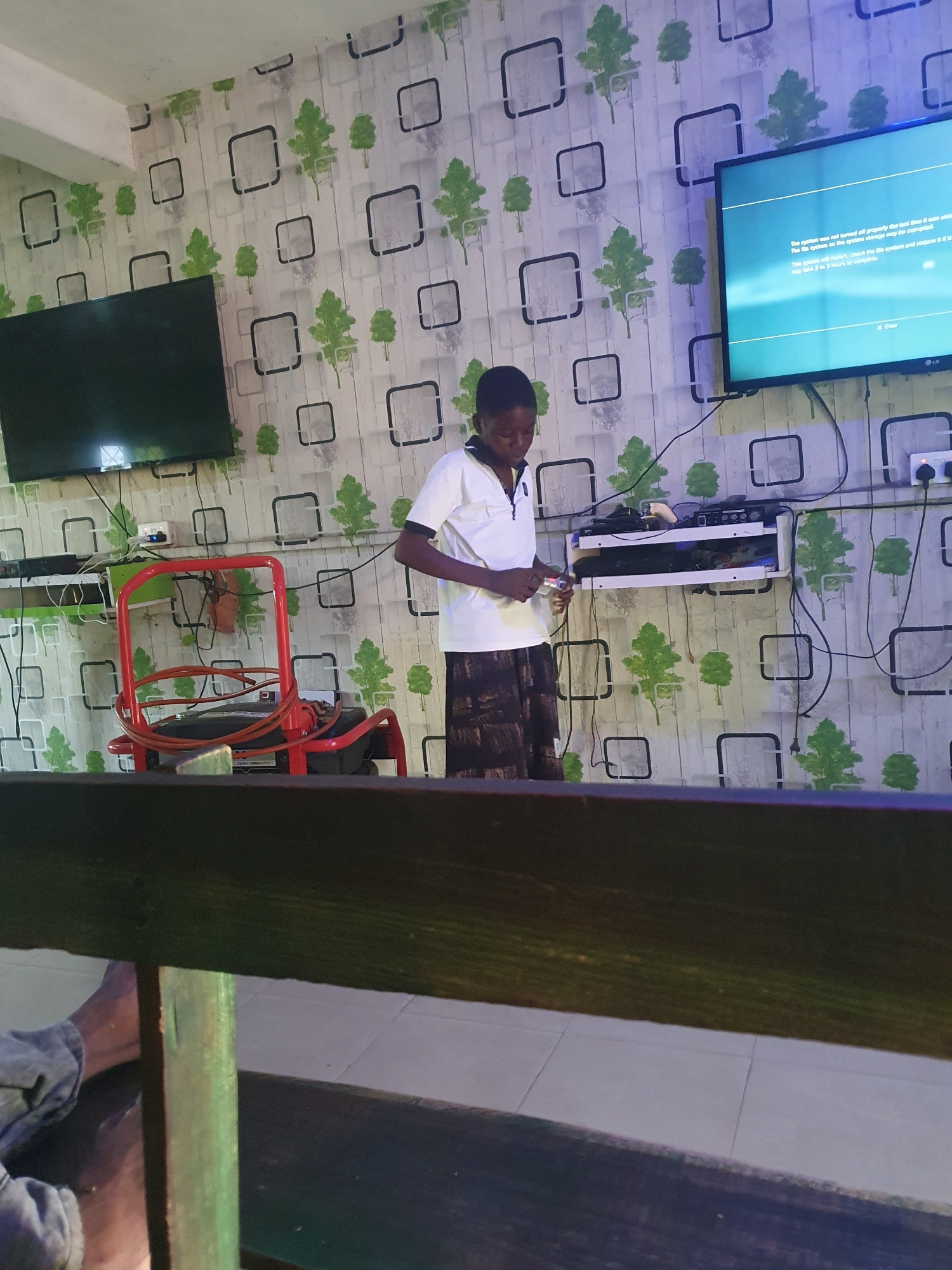 In an indoor setting with patterned wallpaper featuring green trees and grey squares, a young person stands in the foreground, facing right and looking down at their hands. They are wearing a white collared shirt with a zipper and dark patterned trousers. The person appears to be holding a small object.
Behind them, a television screen displays text that reads "The system was not turned off properly the last time it was used. The file system on the system storage may be corrupted. This system will restore, check the file system and restore it. This may take 2 to 3 hours to complete." Below this text is an option to "X Enter."
To the left of the television, a black flat-screen television is mounted on the wall. Below it, a white shelf unit is attached to the wall, holding various electronic devices and cables. A red chair with a generator on it is positioned to the left of the shelf unit.
The environment appears to be a room with tiled flooring visible in the lower portion of the image. A dark wooden beam runs across the bottom of the frame, obscuring the lower parts of the scene.