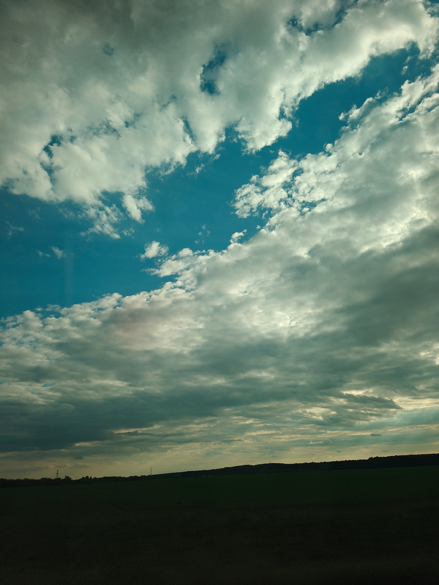 The image depicts a vast, overcast sky with dramatic cloud formations, transitioning from bright, white cumulus clouds towards the top left, to darker, more diffused grey clouds occupying the majority of the upper frame. A sliver of deep blue sky is visible through a gap in the clouds on the left side. The lower portion of the image shows a dark, silhouetted landscape, likely a field, with a faint, dark tree line and the suggestion of distant hills or a horizon. Several utility poles are sparsely visible along the horizon line. The overall impression is of a wide-open, rural or semi-rural environment under a dynamic sky, consistent with an outdoor scene in the vicinity of Zinkiv, Ukraine, given its geography and typical weather patterns. There are no people, animals, or discernible actions taking place.