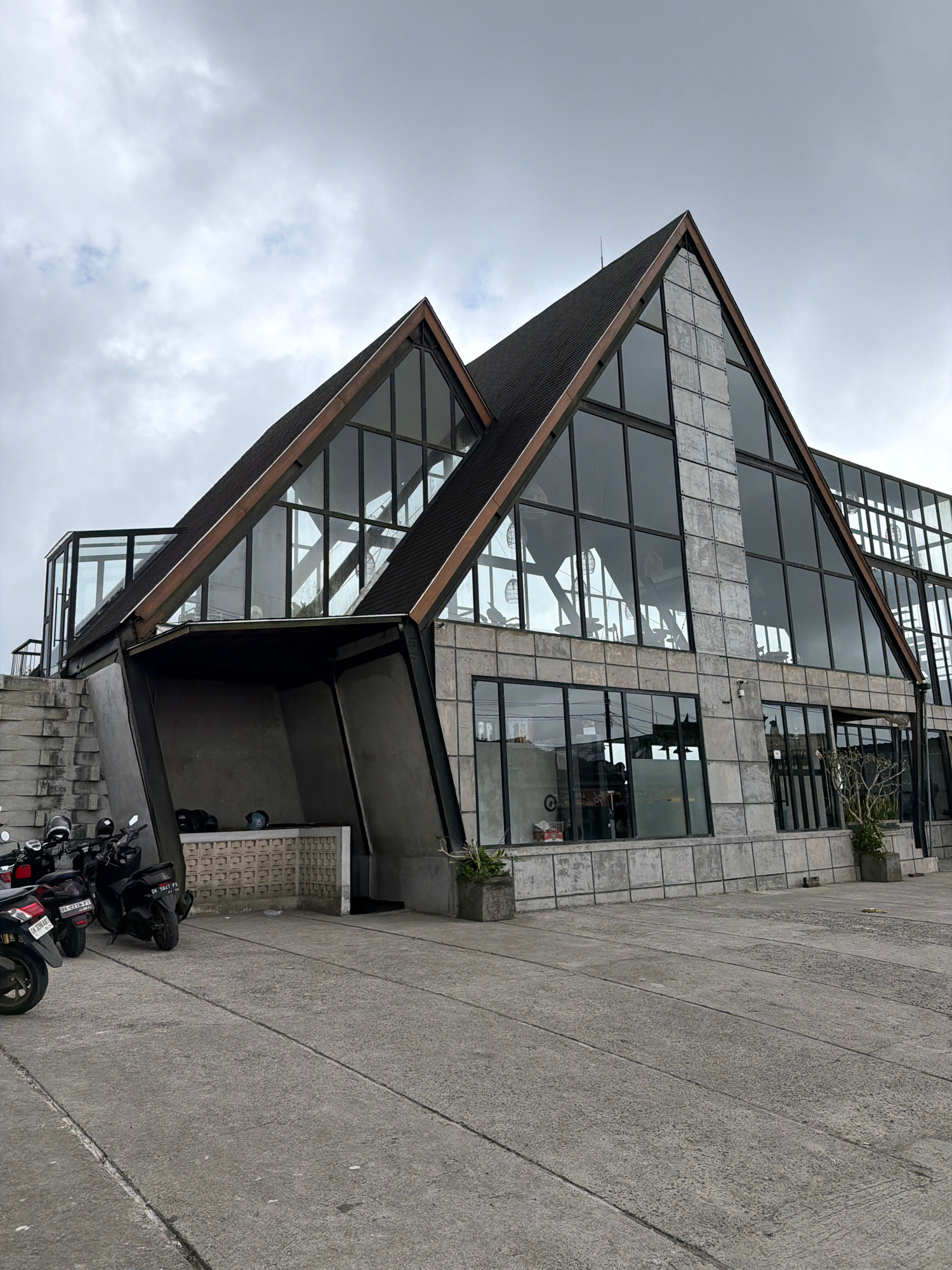 The image depicts a modern building with a distinctive angular roof structure and extensive glass windows, situated in Kintamani, Indonesia. The building's facade is a combination of concrete and glass, with a dark-pitched roof. Several motorcycles are parked in the foreground on a concrete paved area. The sky above is overcast. The building appears to be a commercial establishment or a unique architectural design, possibly a cafe or restaurant, given the context of Kintamani being a tourist destination. No people are clearly visible in the scene.