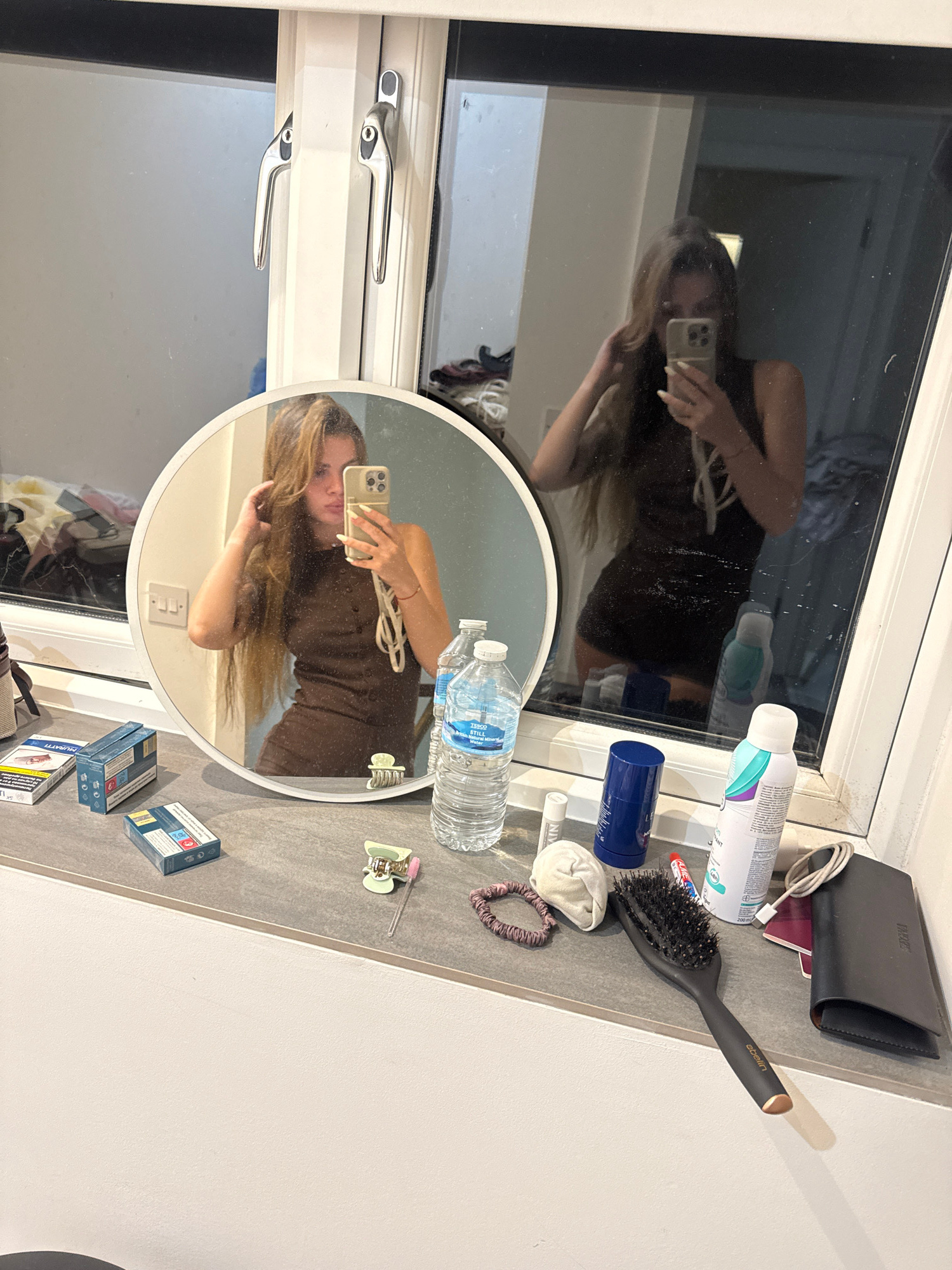 The image depicts a bathroom setting, likely a personal vanity area, with a window in the background. The focus is on a woman taking a mirror selfie, with her reflection visible in a round mirror placed on a countertop. She is wearing a dark, sleeveless dress and has long, light brown hair. She is holding her phone in her right hand and touching her hair with her left.
On the countertop, various objects are arranged, including two packs of cigarettes, a bottle of "TESCO STILL Natural Mineral Water," a blue deodorant stick, a can of hairspray, a black hairbrush with a gold accent, a passport holder, a charging cable, a small white cloth, and several hair clips. The environment appears to be tidy, with the window reflecting the interior. The city of Throop, United Kingdom, is mentioned as the location, although there are no specific visual cues in the image to confirm this location.