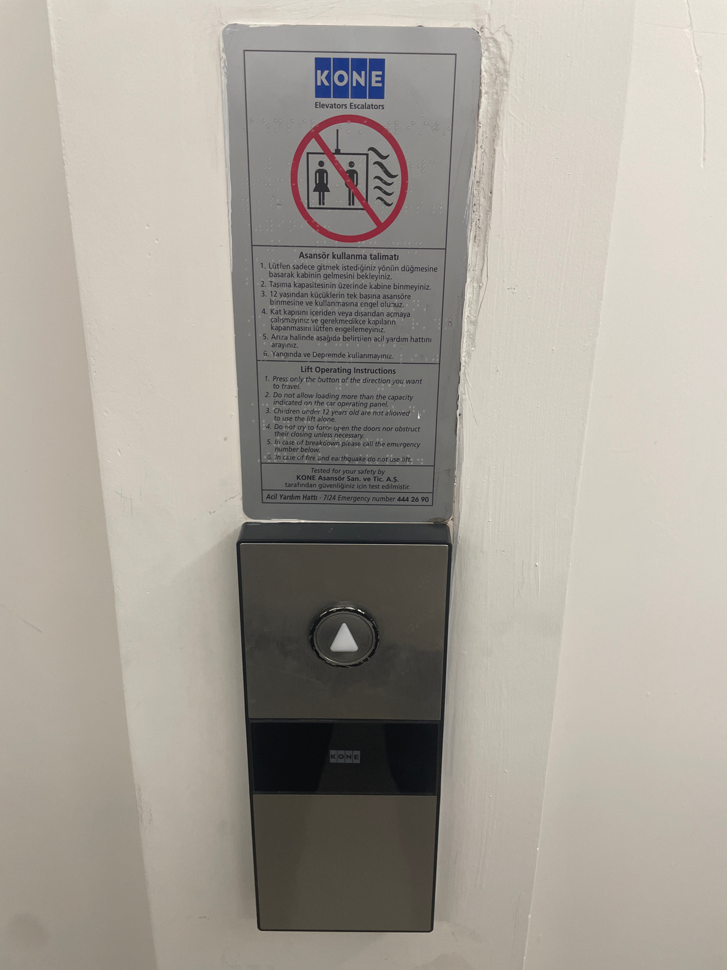 The image shows an elevator control panel with a sign providing operating instructions in both Turkish and English. The sign features the KONE logo and an illustration indicating safety precautions for elevator use. Below the sign, the control panel includes a lit triangular button for up, and a black section with the KONE logo. The surrounding environment appears to be an indoor wall, possibly in a building in Yunus Emre, Turkey, given the presence of Turkish language on the sign. The wall is textured and painted white, with some minor imperfections. The overall scene is well-lit, suggesting it's an interior space.