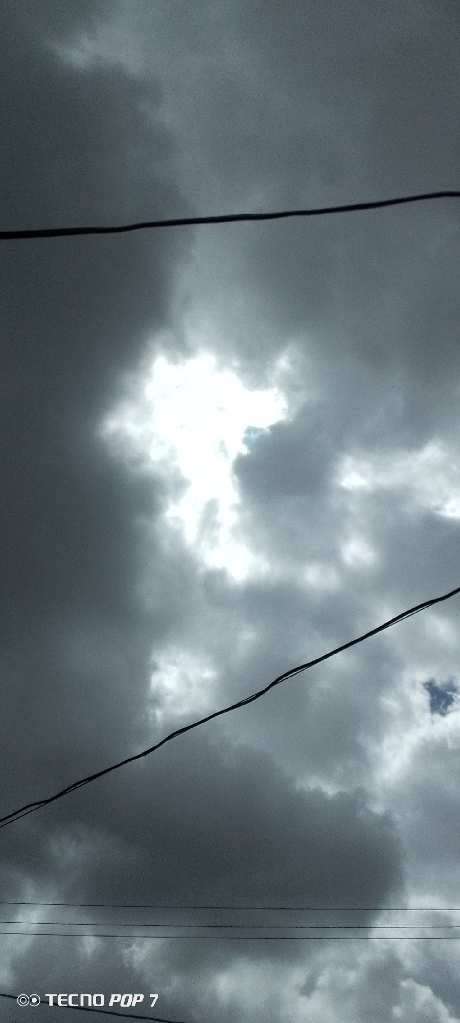 The image captures a segment of the sky above Altagracia, Venezuela, characterized by a dramatic interplay of dark, heavy clouds and a bright, almost blinding patch of sunlight breaking through. Multiple power lines are visible crisscrossing the frame, indicating an urban or semi-urban setting. The overall tone is somber and moody, with the stark contrast between light and shadow dominating the scene. No people or specific structures are discernible, focusing the composition solely on the atmospheric conditions and the presence of utility infrastructure. The bottom left corner of the image displays the text "TECNO POP 7," a watermark likely indicating the device used to capture the photograph.