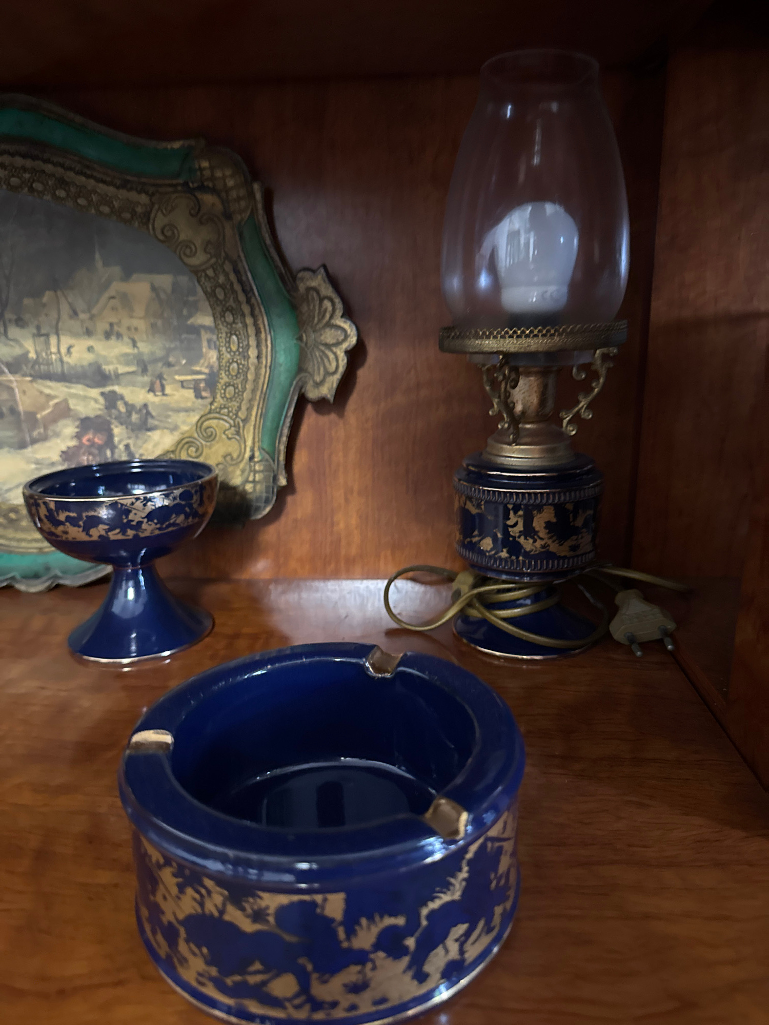 The image shows a close-up view of decorative objects placed on a wooden shelf. In the foreground, there is a dark blue ceramic ashtray with gold detailing. To the left and slightly behind the ashtray, there is a smaller, dark blue ceramic cup with a pedestal base, also featuring gold accents. Further back and to the left, a framed decorative plate with a painted winter scene is visible. On the right side of the image, a small, ornate table lamp with a glass shade and a blue and gold base is present. The lamp has a cord and plug. The objects appear to be arranged inside a wooden cabinet or bookshelf, with the wood grain of the shelf and the cabinet walls visible. The lighting is somewhat dim, suggesting an indoor setting. No people are visible in the image. The location in Gandia, Spain is not discernible from the visual information provided.
