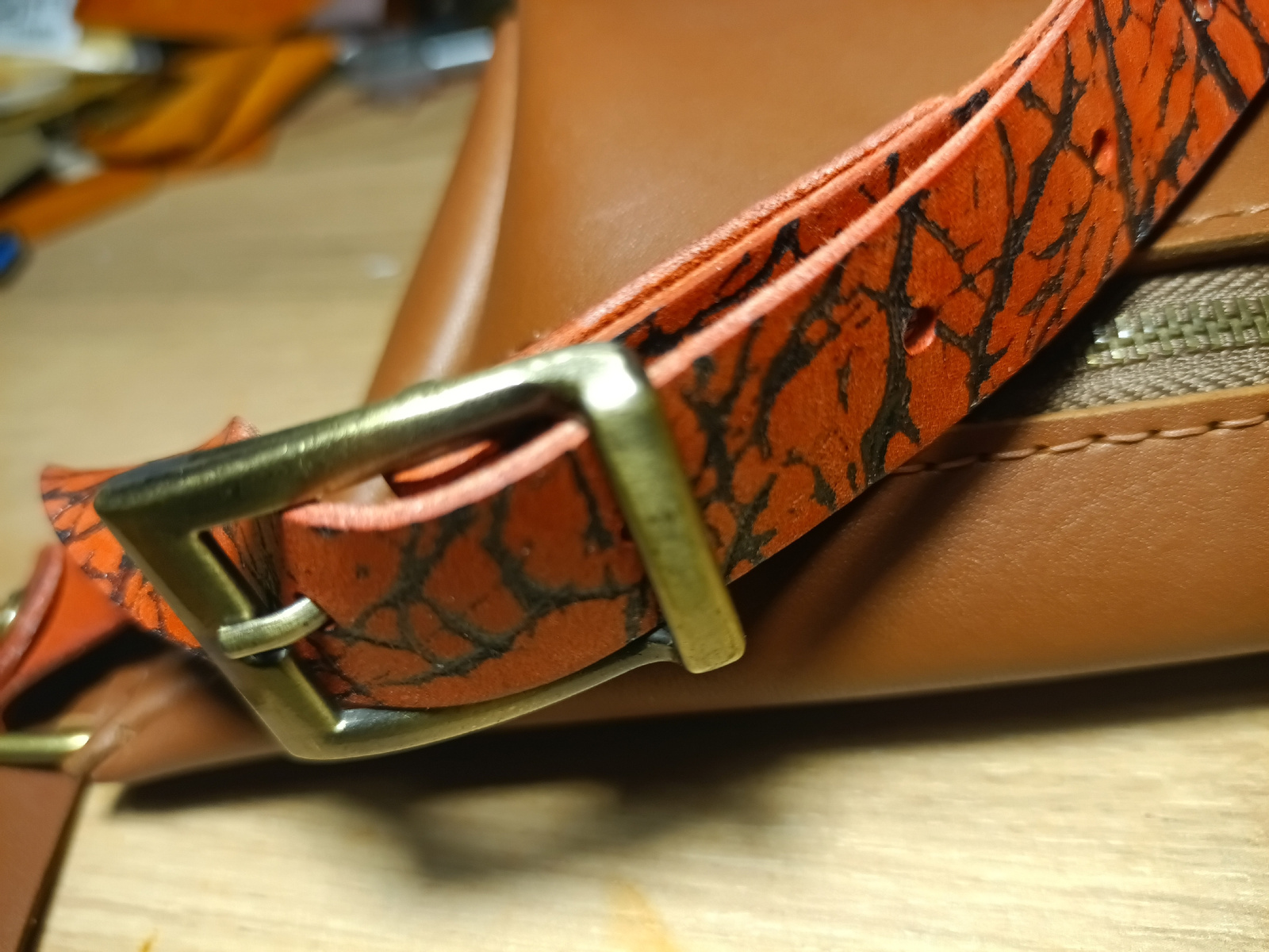 This is a close-up, low-angle, eye-level shot of a patterned leather strap attached to a metal buckle. The strap is orange with a black, cracked pattern that resembles tree bark or lightning strikes. It is threaded through a rectangular, antique brass-colored buckle. The strap appears to be part of a bag or accessory, as a portion of a smooth, brown leather surface is visible behind it, along with the teeth of a zipper. The background is blurred, showing a light-colored wooden surface, possibly a table or countertop. The lighting is somewhat dramatic, casting shadows and highlighting the textures of the leather and metal. There is no visible text. The overall impression is of a detail of a handcrafted item, emphasizing its material and design. Given the lack of human presence and clear context, the specific activity is unknown, but it appears to be a static display or examination of the object. The setting is likely an indoor environment, possibly a workshop or a retail display.