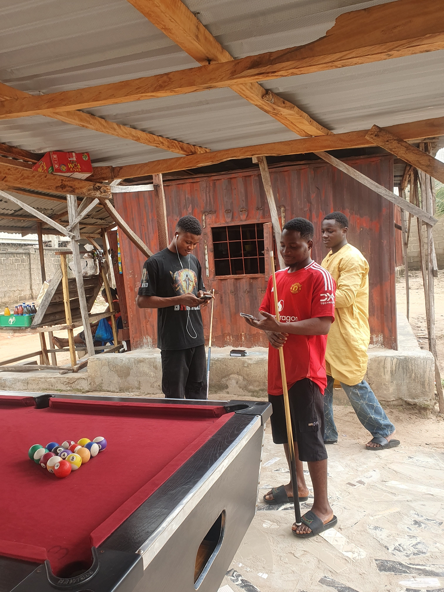 Three young Black men are gathered around a red pool table under a makeshift wooden shelter in Lokoja, Nigeria. The shelter has a corrugated metal roof and wooden beams. One man in a black t-shirt and black shorts is looking at his phone while holding a pool cue. Another man in a red Manchester United jersey and black shorts is also looking at his phone and holding a pool cue. The third man, wearing a yellow traditional Nigerian tunic and blue jeans, stands behind them. The pool table is set up with a triangle of pool balls. The ground is a mixture of concrete and dirt. In the background, there is a rustic, corrugated metal structure that appears to be a building or shed, with a fenced area and some greenery visible beyond. Bottles of drinks are stacked on a shelf in the background. The overall scene suggests a casual outdoor gathering place, possibly a bar or recreational area.