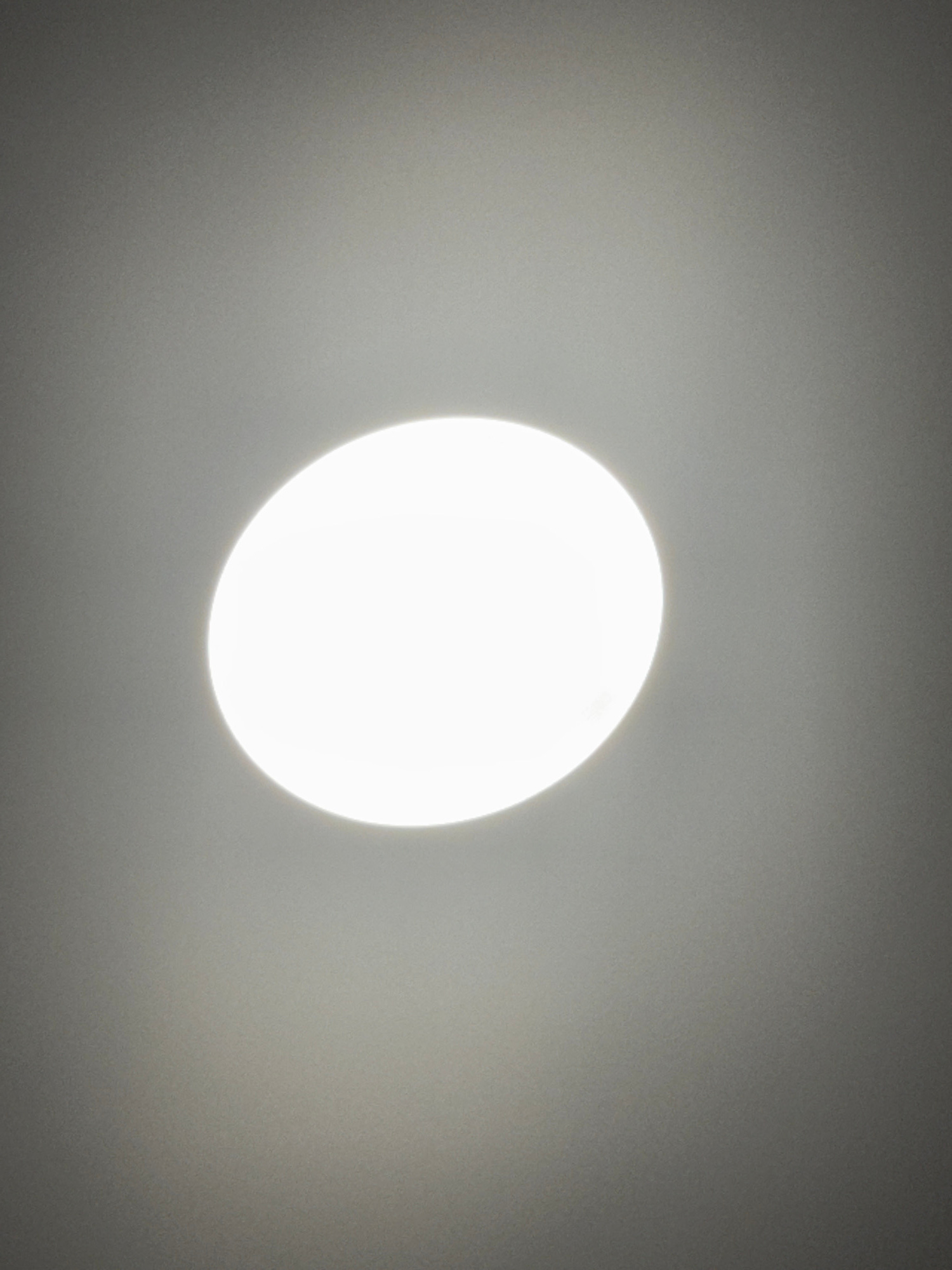 A close-up, overhead shot captures a bright, circular LED ceiling light illuminating a muted grey surface. The light is centered in the frame, its intense glow creating a soft halo effect that fades into the surrounding grey. The surface appears to be a ceiling or a similar flat, opaque material. There are no other objects or people visible in the image. The lighting is flat and even, with no distinct shadows. The image is taken from a low angle, looking directly up at the light fixture. This image does not contain any recognizable landmarks or details that would allow for its location to be identified in Bangkok, Thailand.