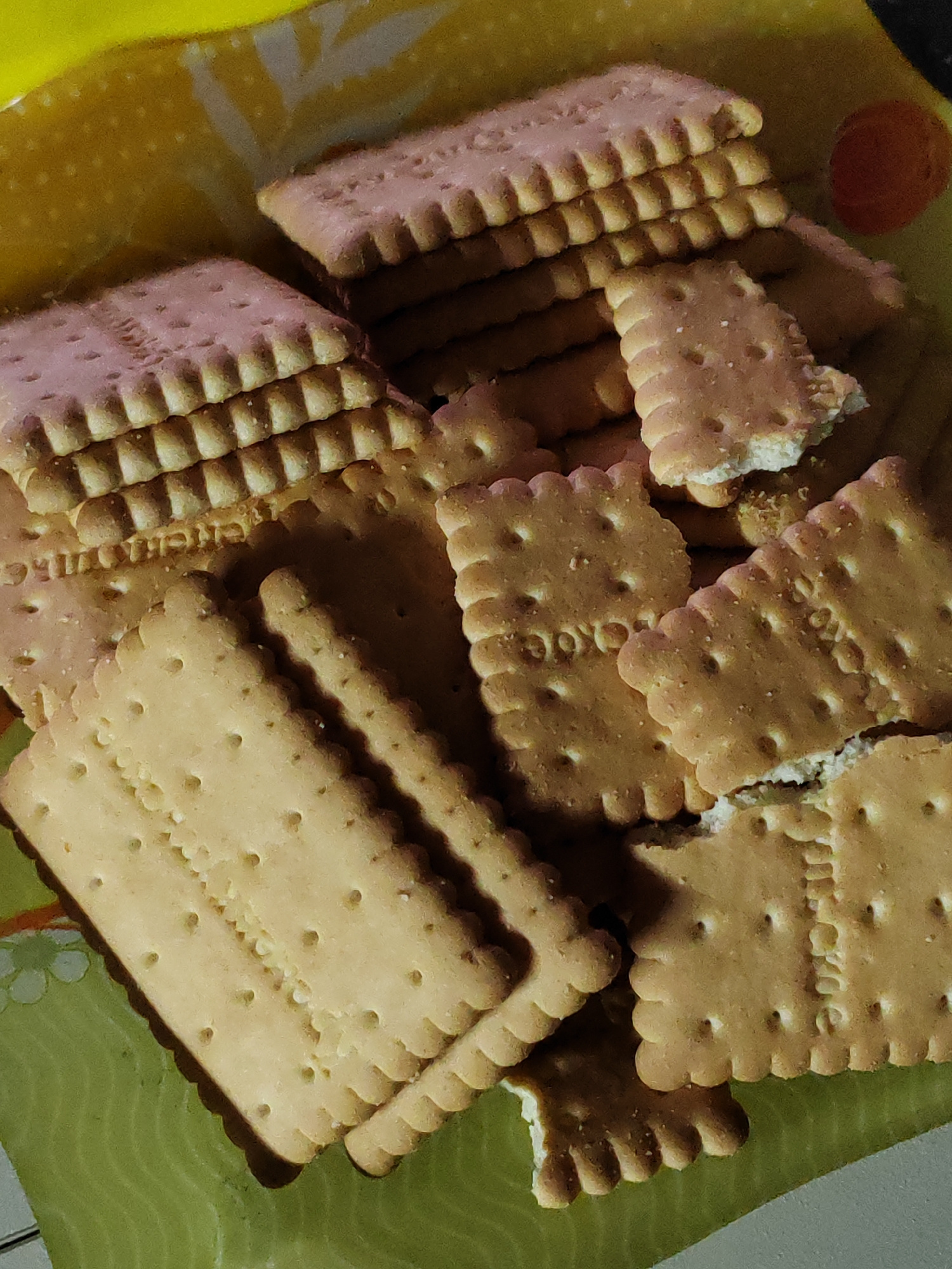 A pile of rectangular, golden-brown biscuits with embossed patterns and serrated edges are arranged on a yellow and green patterned surface. Some biscuits are stacked in neat layers, while others are haphazardly placed, with a few showing signs of being broken. The lighting suggests an indoor setting, possibly a kitchen or dining area, with soft shadows indicating the direction of light. There are no people or animals visible in the image. The scene appears to be a close-up shot of a snack. The location of Unknown, Russia is not discernible from the provided image.