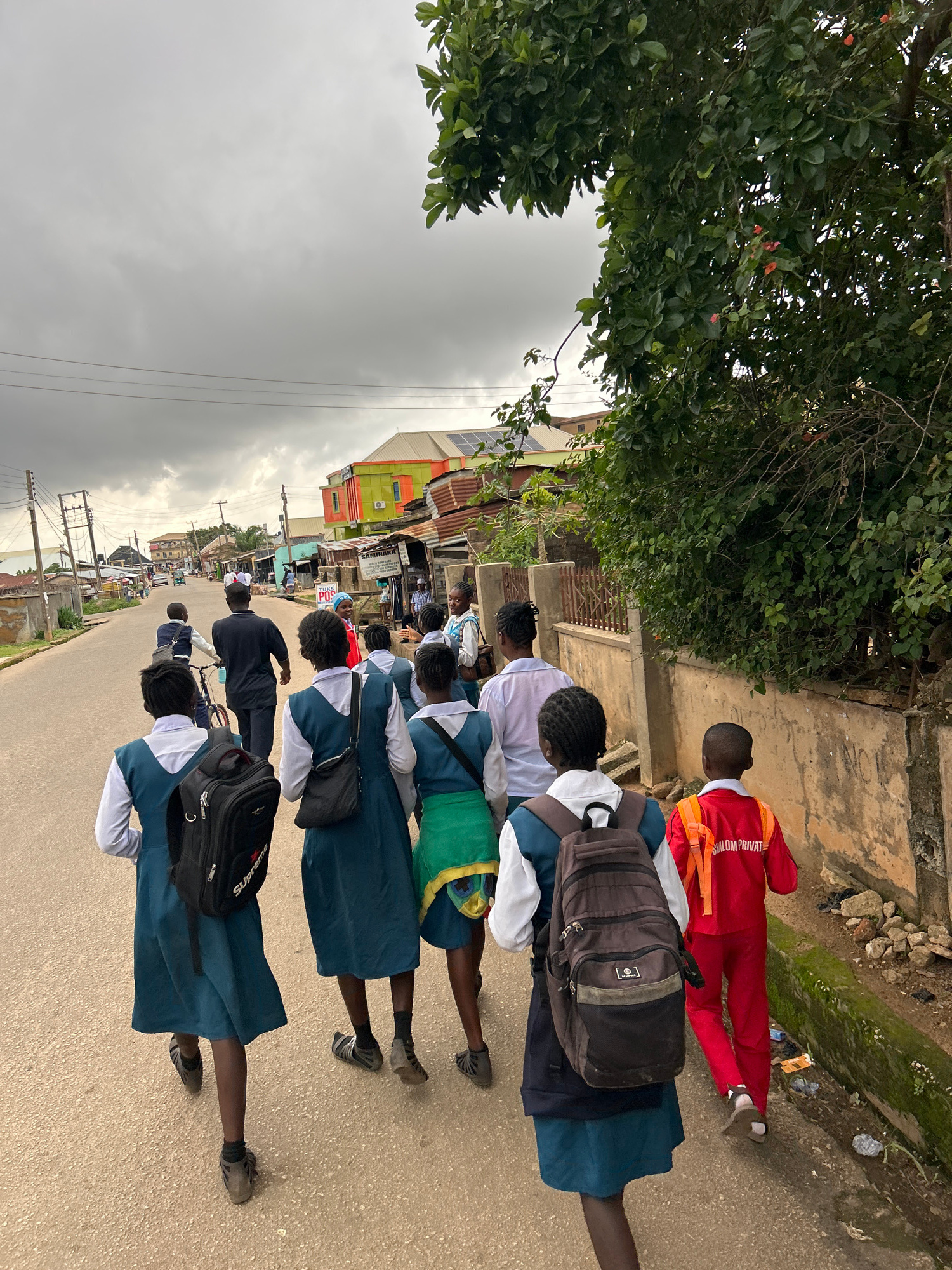 The image shows a group of young people, likely students, walking down a paved street in what appears to be a town or city in Jos, Nigeria. Most of the individuals are wearing uniforms, consisting of white shirts and blue or dark blue skirts or shorts. Some have backpacks, suggesting they are going to or from school. One person in the foreground wears a red uniform with orange straps, which is possibly a different school's uniform or athletic wear.
The scene is set outdoors, with buildings visible on the left side of the street, some with corrugated iron roofs. Lush green trees overhang the scene from the left, partially obscuring the sky. The sky is overcast, with gray clouds suggesting it might be raining or has recently rained. The ground is paved, and there is a drainage channel running alongside the street, which appears to have some green moss and debris.
The people are walking in a line, facing away from the camera, with the exception of the person in red who is facing sideways. Their expressions are not visible, but their postures suggest they are on the move, perhaps in an orderly fashion. The overall impression is one of daily life, possibly the end of a school day or the beginning of one.
Visible text includes "SHALOM PRIVATE" on the back of the red uniform. Another backpack has a logo that appears to say "SUPREME". There are also signs on some of the buildings, but they are not legible in detail. The time of day appears to be daytime, given the ambient light, but the cloudy weather makes it difficult to pinpoint more precisely.