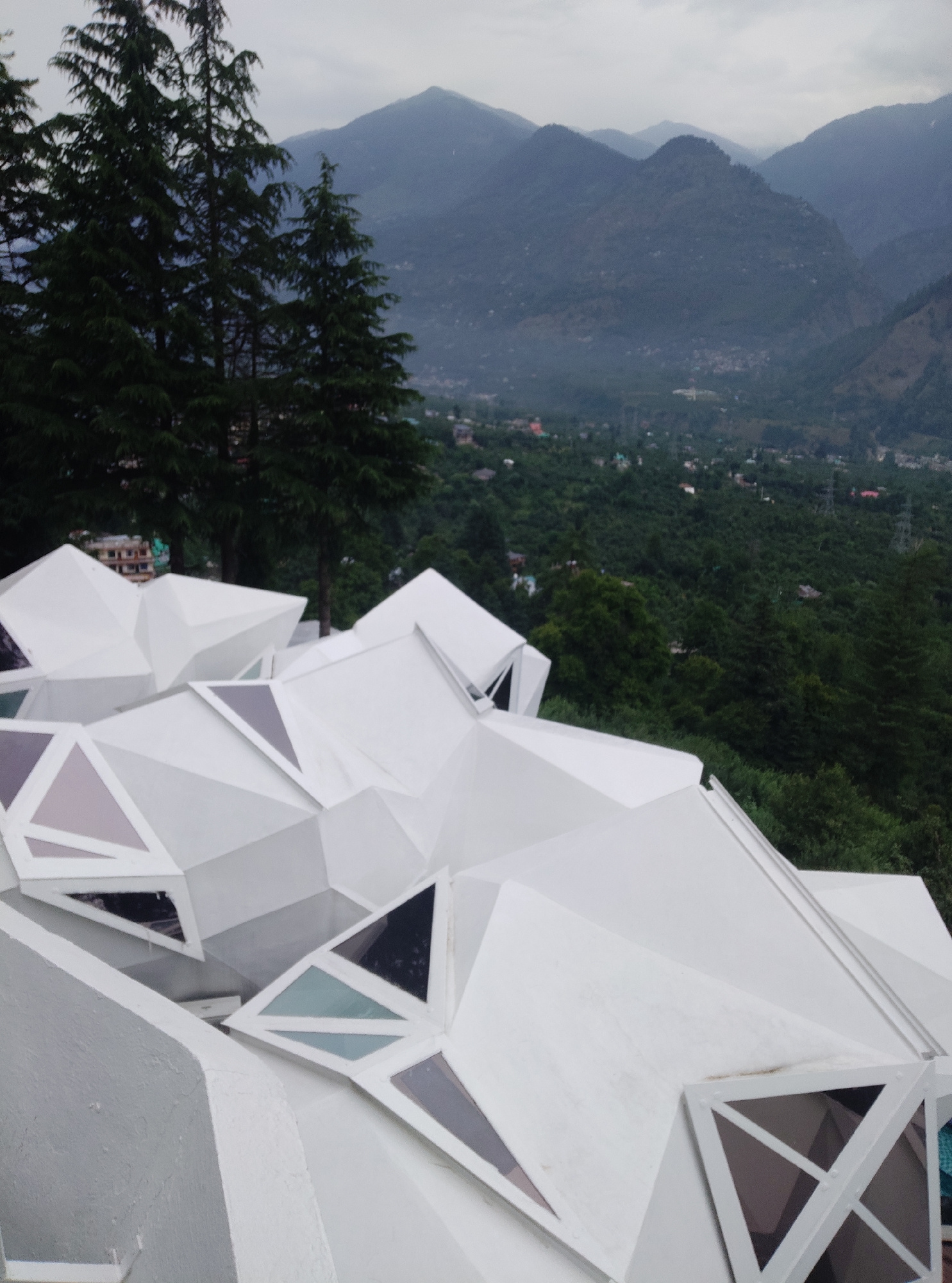 The image depicts a modern, angular white structure situated on a hillside overlooking a valley. The structure is composed of numerous sharp, geometric facets and features triangular windows with varied colored panes, some opaque and some translucent. In the background, lush green hills rise, densely covered with trees, and a range of larger, misty mountains is visible on the horizon. The sky is overcast and grey. Tall, dark green coniferous trees are prominent in the foreground to the left, partially obscuring the view of the valley below. The overall scene suggests an architectural design integrated into a natural, mountainous environment.
