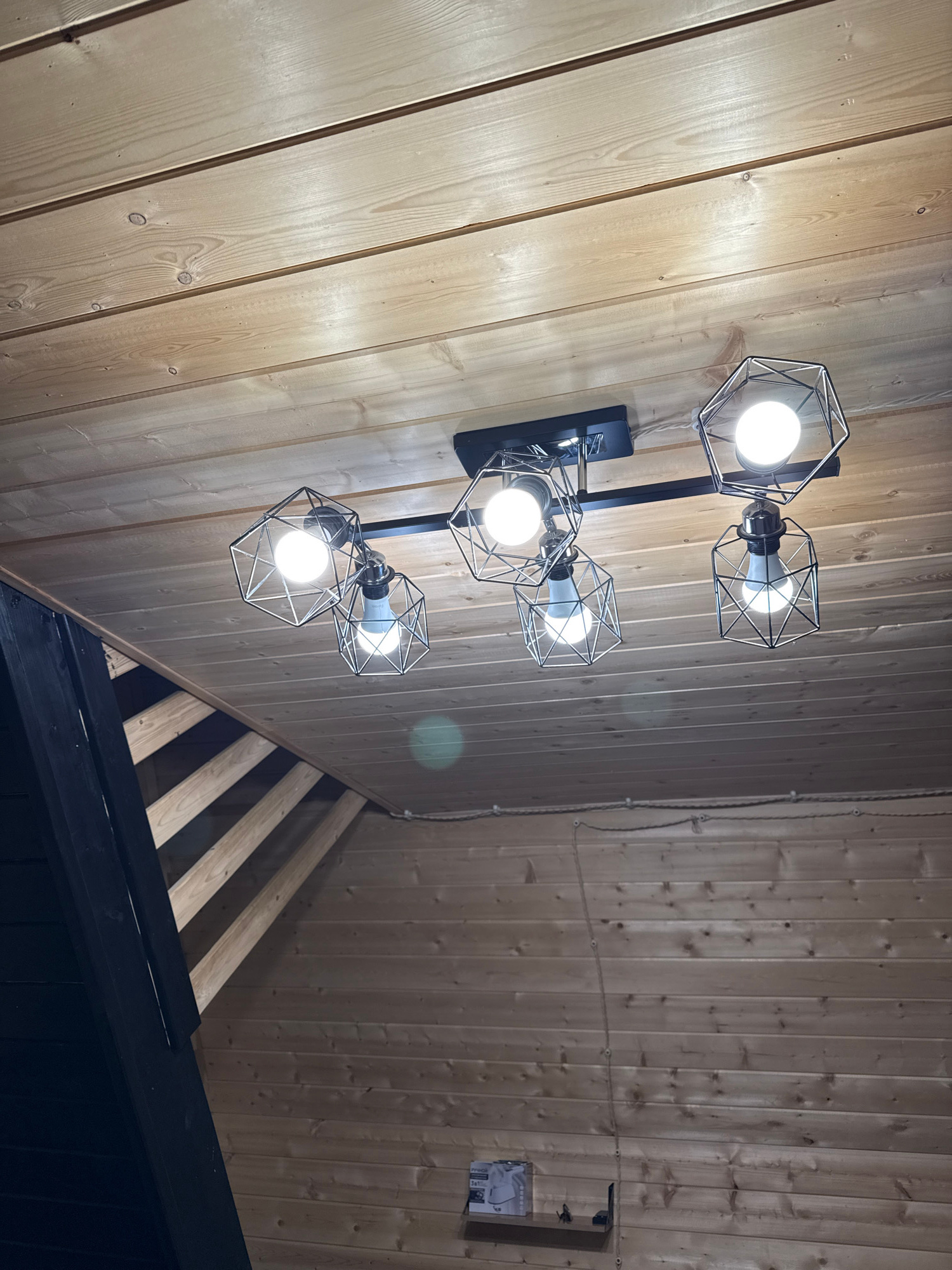The image shows a modern light fixture with a black base and five hexagonal wire cages, each containing an LED bulb, mounted on a wooden plank ceiling. The ceiling planks run horizontally across the frame. To the left, a dark-colored structure, likely a wall or roofline, slopes upwards, revealing wooden beams underneath. To the right, more wooden planks form a sloped ceiling or wall. A thin rope with visible fasteners is strung horizontally across the upper portion of the right side. In the lower right quadrant, a light-colored wooden shelf is mounted on the wall, holding a small box and what appears to be a decorative object. The overall environment suggests an interior space with a rustic or cabin-like aesthetic, possibly in a residential or recreational building in Arkhyz, Russia, given the presence of wood and the sloped roofline which is common in mountainous regions. There are no people or animals visible in the image.