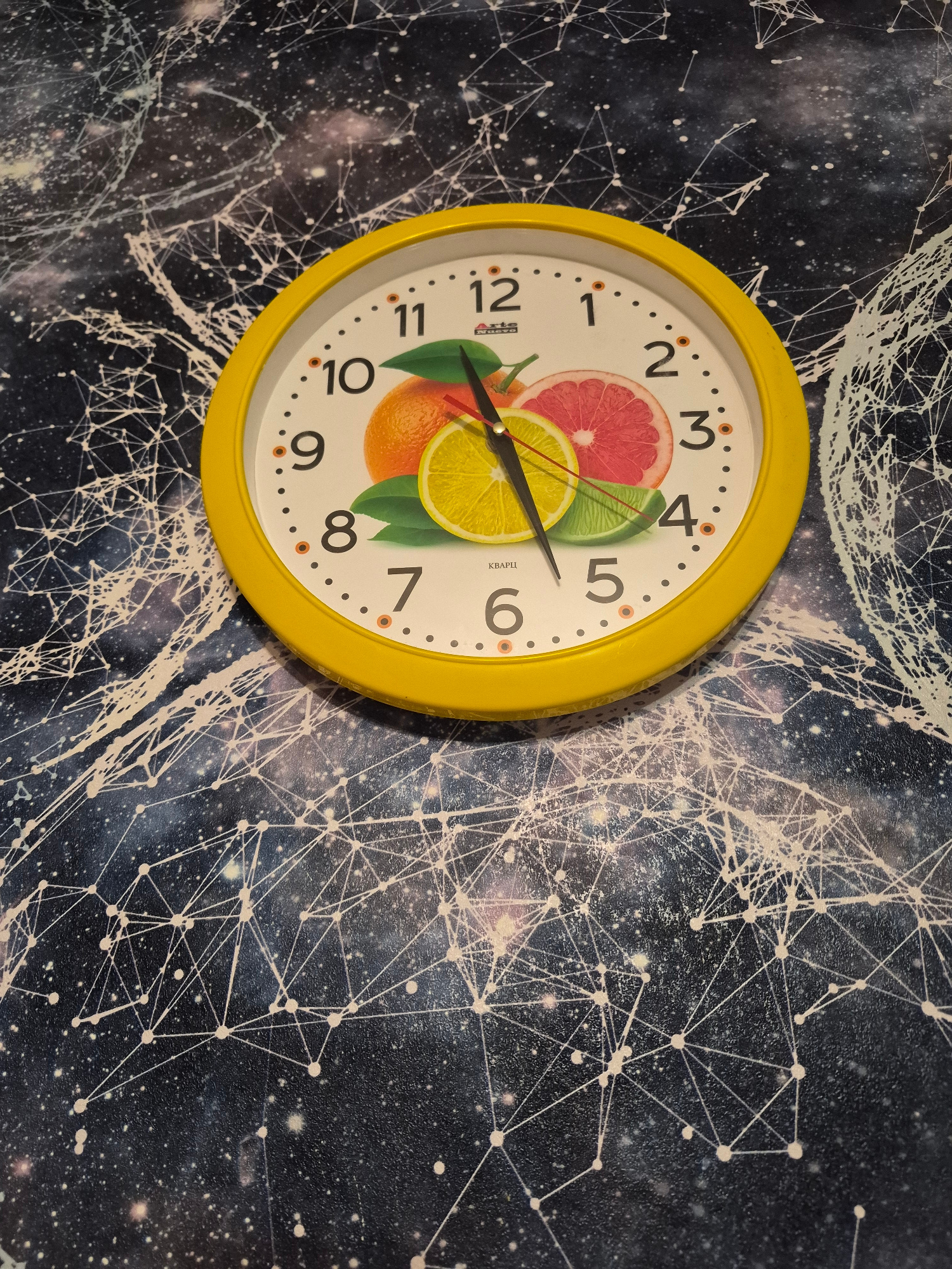 A yellow-framed analog clock is displayed on a dark surface with a white constellation pattern. The clock face features images of citrus fruits, including an orange, a grapefruit slice, and a lemon slice. The clock hands are black. The hour hand points between the 2 and the 3, and the minute hand points between the 10 and the 11. The time displayed is approximately 2:53. The location in Nyagan, Russia is not specified in the image.