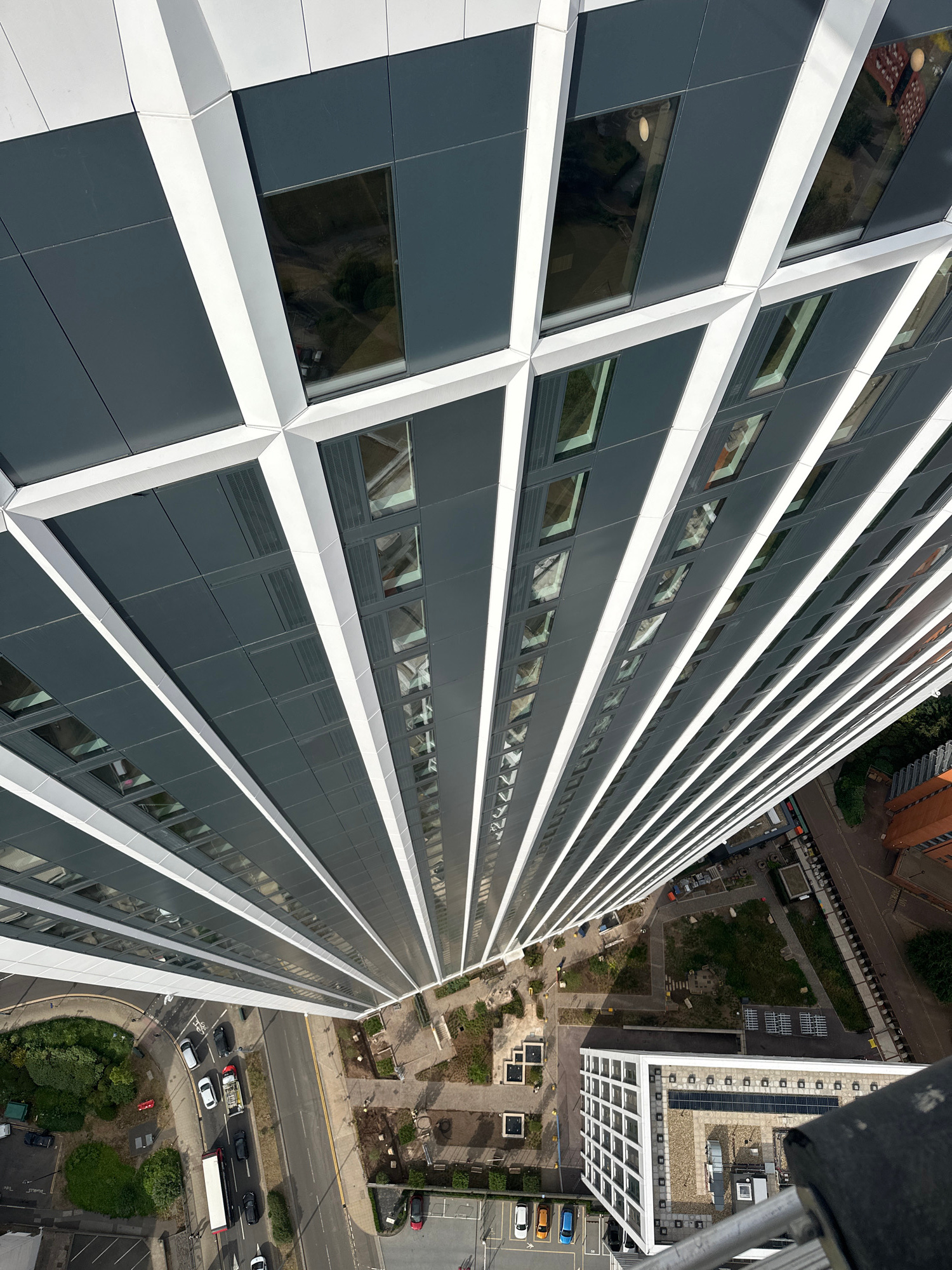The image provides an extreme high-angle, aerial perspective looking down the side of a modern, multi-story building in Birmingham, United Kingdom. The building features a distinctive facade composed of dark grey panels and white vertical and diagonal accents, creating a geometric pattern. Numerous windows are visible, reflecting the sky and surrounding environment.
Below the building, a street with traffic is visible, including cars, a bus, and a fire truck. The street curves around a green area with trees and landscaping. Further down, another building with a white grid facade and solar panels on its roof can be seen. The scene appears to be a daytime urban environment, possibly a commercial or residential development. There are no visible people in the immediate foreground or detailed enough in the background to identify. The overall impression is one of contemporary urban architecture and infrastructure.