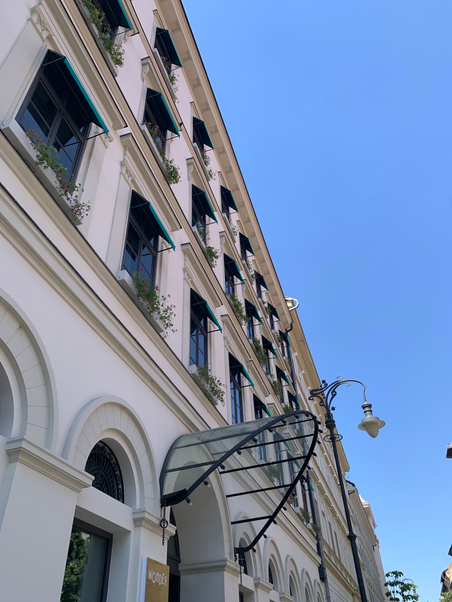 The image captures a low-angle view of a historic building facade against a clear blue sky. The building is white with multiple stories, featuring numerous windows each adorned with a dark green awning and small potted plants. Decorative molding is visible around the windows and along the building's edges. An arched entrance with intricate black metalwork above is prominent on the left side, and a modern glass canopy extends over another entrance. A vintage-style street lamp with an ornate black metal arm stands to the right of the building, its light fixture a creamy white. The overall impression is one of architectural elegance and historical character. Based on the architectural style and the presence of the "WONDER" sign which could be associated with a boutique or hotel, this scene is likely located in Budapest, Hungary.