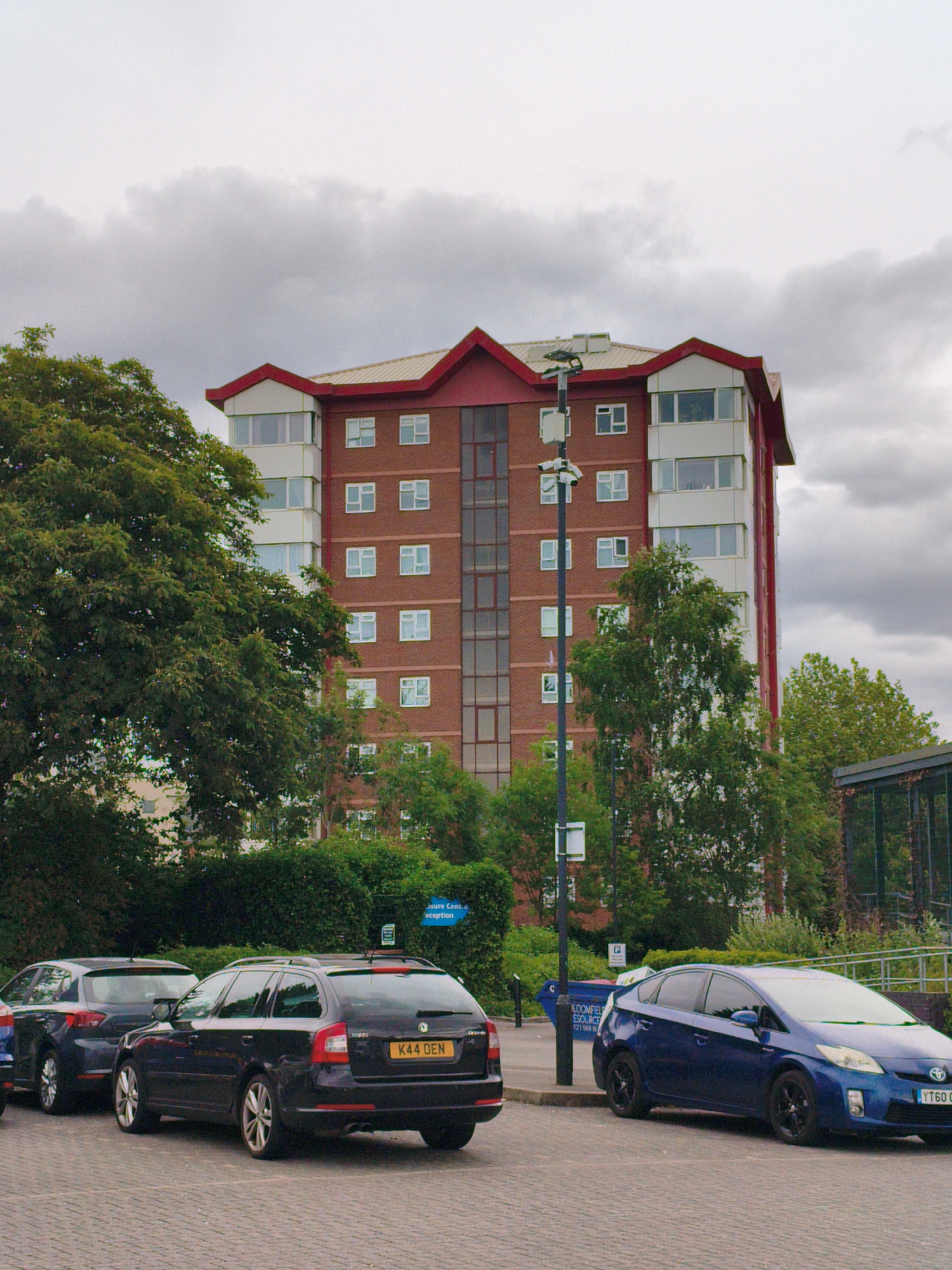 This media file shows a street scene with a tall brick building with a red roof. A streetlamp with security cameras is visible in front of the building. Several cars are parked in a paved lot in the foreground, including a dark blue Skoda estate with the license plate "K44 DEN" and a blue Toyota Prius with the license plate "YT60 O__". Trees and bushes surround the building and parking lot. The sky is overcast, suggesting it is a cloudy day. The location is identified as Sandwell, United Kingdom. There is a blue dumpster with text that appears to read "BLOOMFIELD RESOURCE" and a sign in the background with blue text that reads "Leisure Centre Reception".
