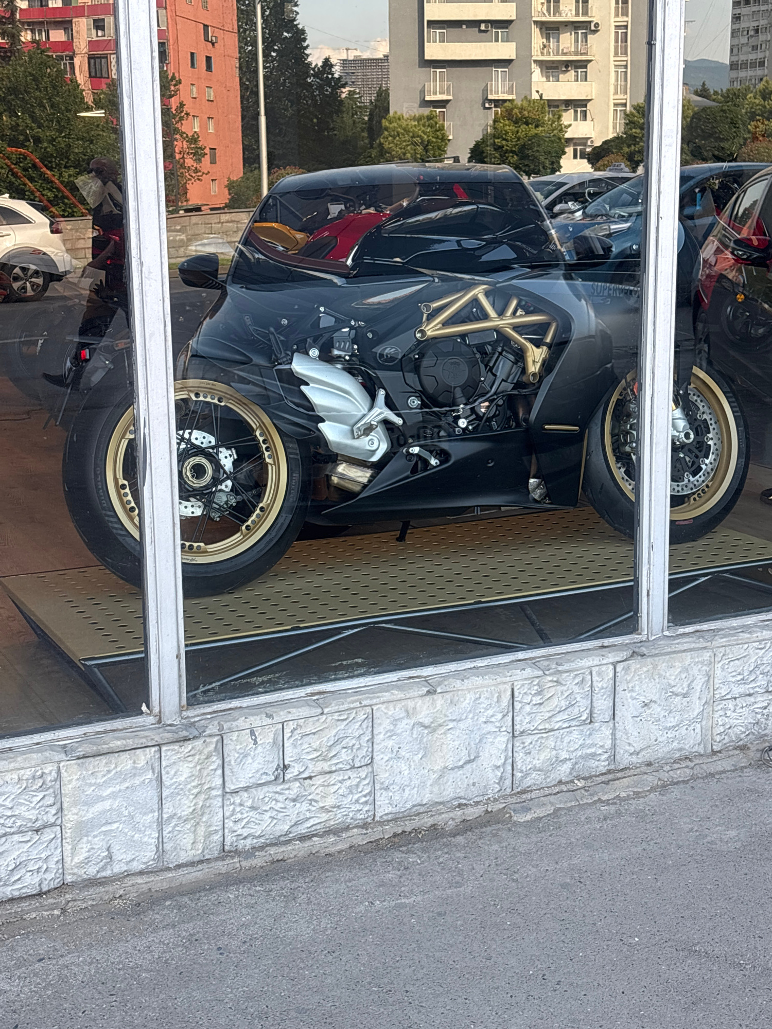 The image shows a motorcycle displayed behind a large window, reflecting buildings and trees in the background. The motorcycle is black with gold accents, and its engine is visible through the bodywork. It is positioned on a textured yellow platform. The window is framed by white vertical elements and a stone-patterned wall below. Reflections in the window show buildings with balconies and trees, as well as other vehicles. The scene is outdoors, as indicated by the lighting and the presence of trees and buildings typical of an urban environment. Based on the architectural style and the presence of a white sedan that appears to be a Peugeot 208, the location is identified as Tbilisi, Georgia, specifically on Griboedov Street, near the intersection with Gamsakhurdia Avenue, and adjacent to a building with distinctive red brick detailing.