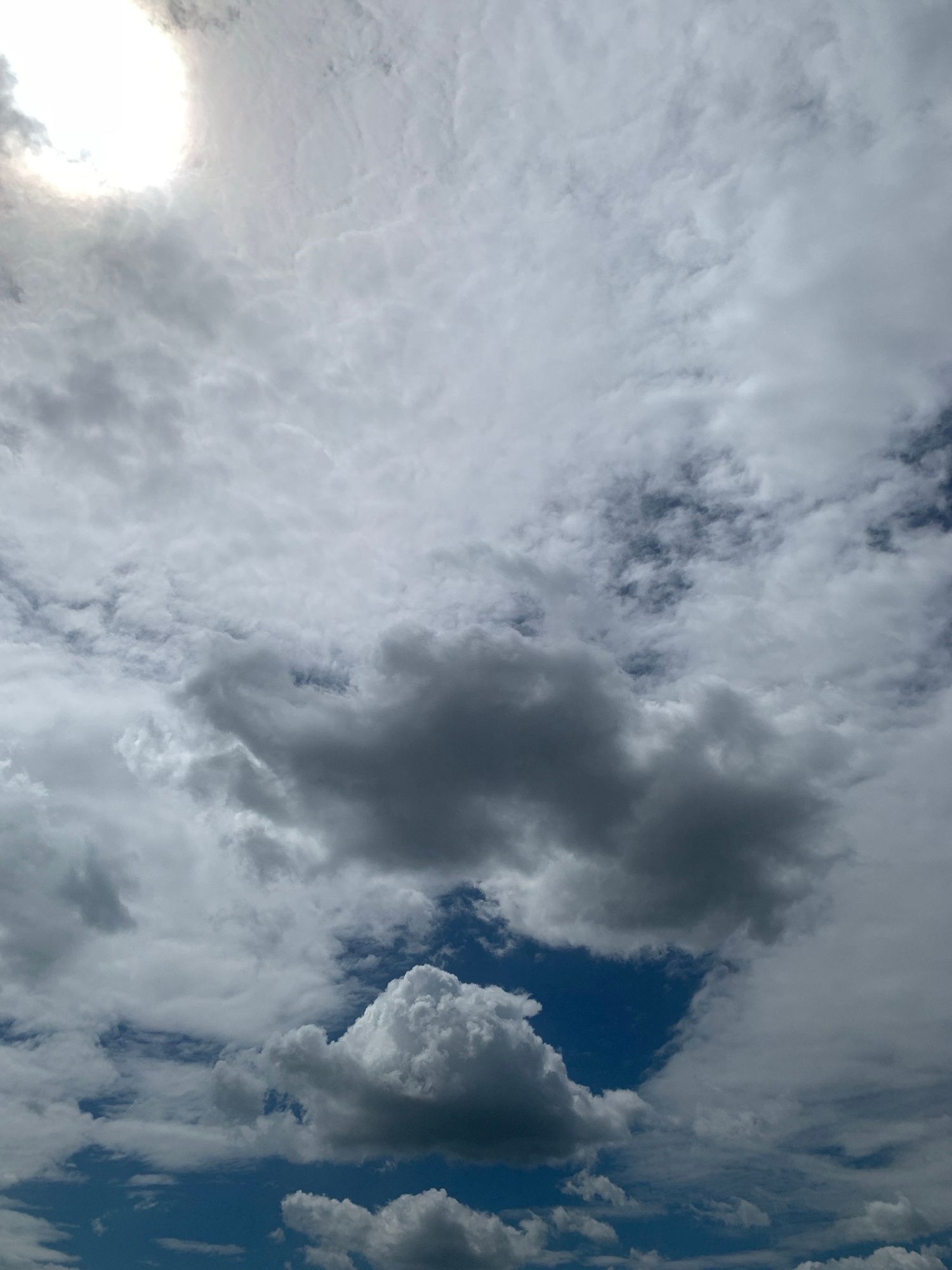 The image shows a sky filled with dramatic clouds and a bright sun in the upper left corner. The clouds are a mix of bright white and dark grey, suggesting a partly cloudy or stormy day. The blue sky is visible in patches between the clouds. The scene is a natural landscape without any visible urban elements or people.