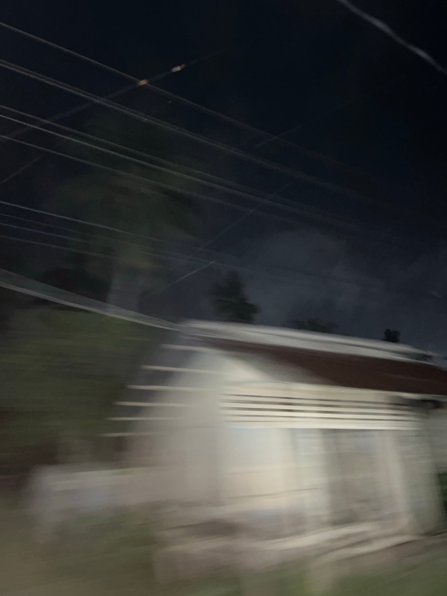 An overexposed, motion-blurred nighttime outdoor photograph shows a blurry, indistinct residential structure with horizontal siding and a darker roof. A network of utility lines traverses the dark sky above the building. The overall impression is one of motion, possibly captured from a moving vehicle, obscuring fine details of the building and its surroundings. No people or specific identifiable landmarks are visible. The location in Buleleng, Indonesia is not discernible from the provided image.